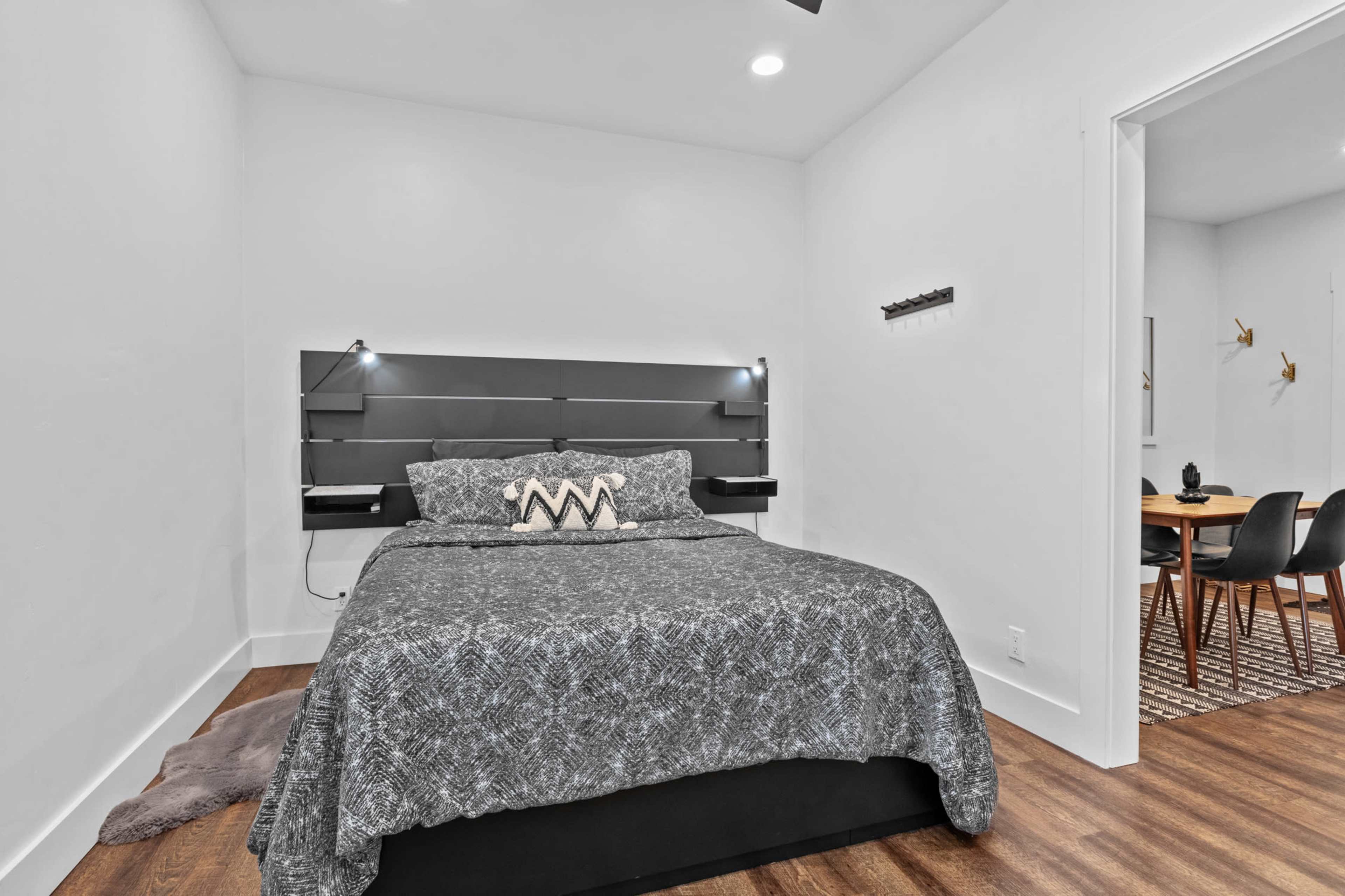 A simple bedroom features a bed with a patterned gray blanket and bedside lamps, along with a dining area visible through an open doorway.