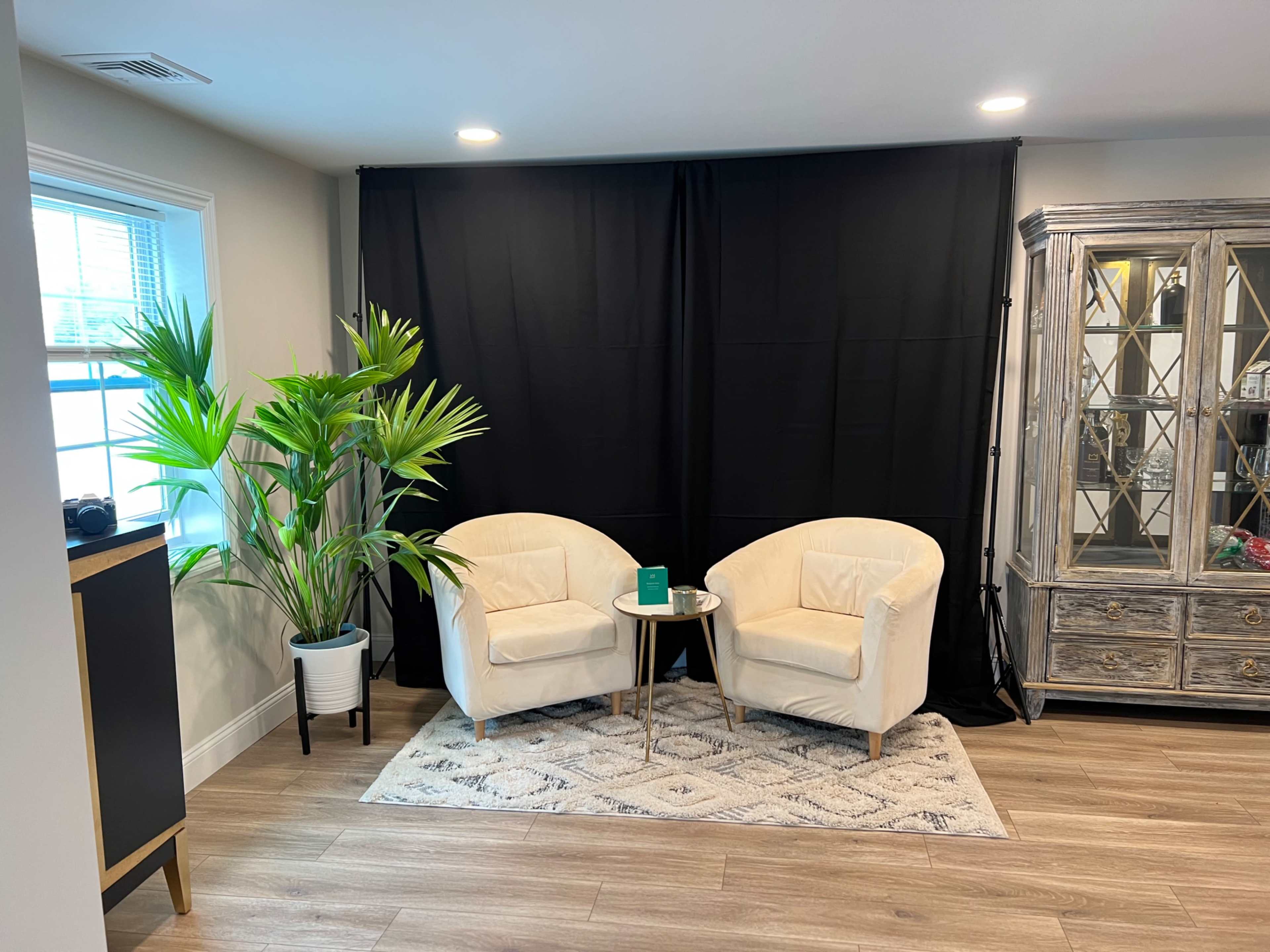 A cozy seating area features two white armchairs, a small round table, and a tall potted plant in front of a black backdrop.