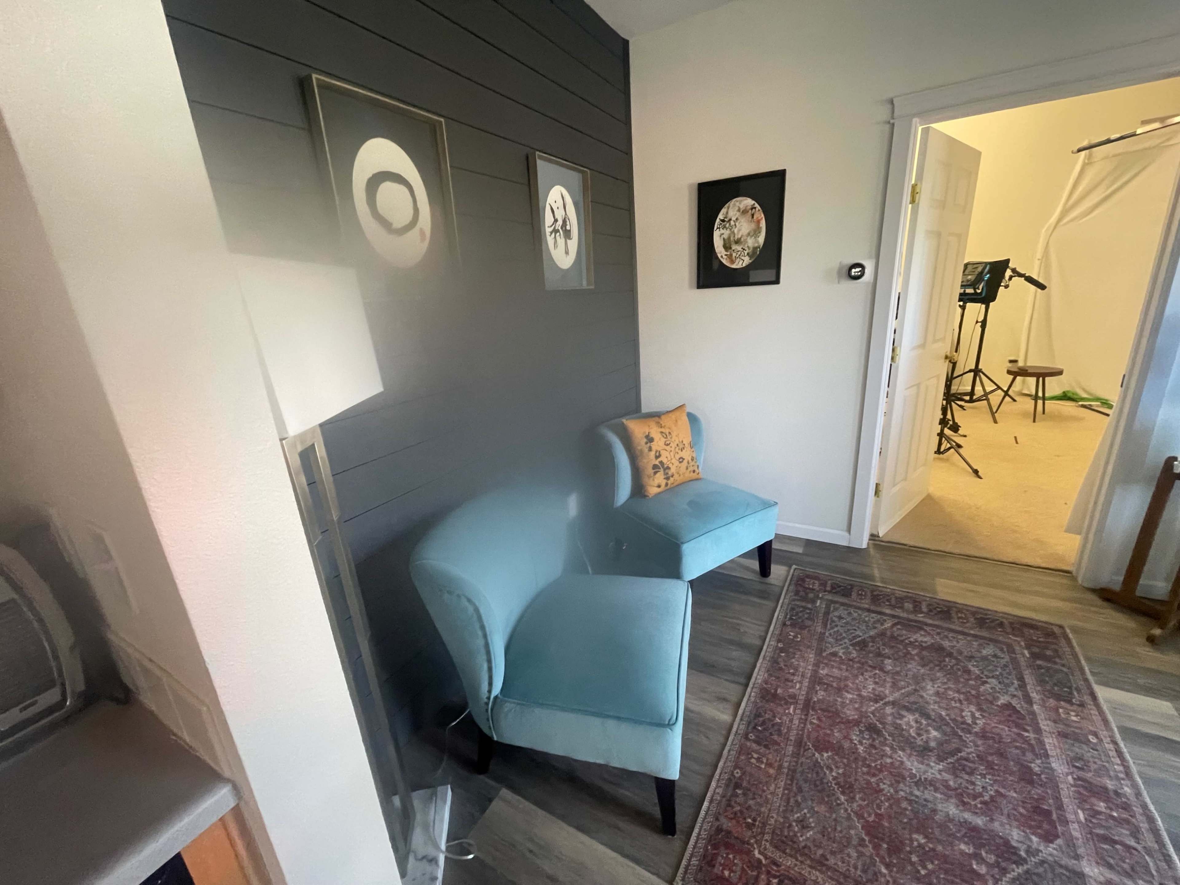 A cozy corner of a room with a blue upholstered chair, a decorative pillow, framed artwork on the wall, and an open door leading to another space.