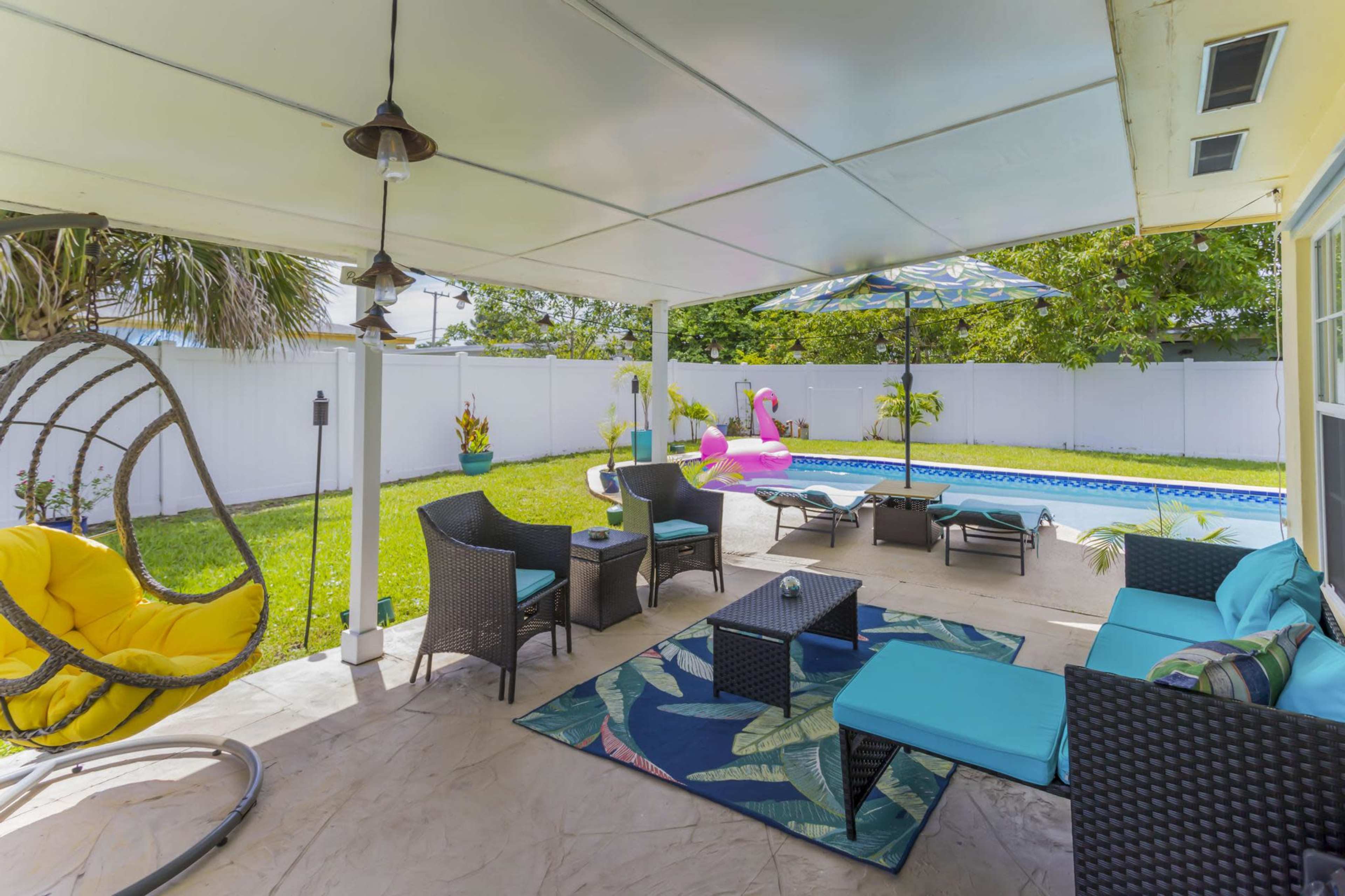 A covered outdoor patio features seating areas, a tropical-themed rug, a swing chair, and a swimming pool with a pink inflatable flamingo.