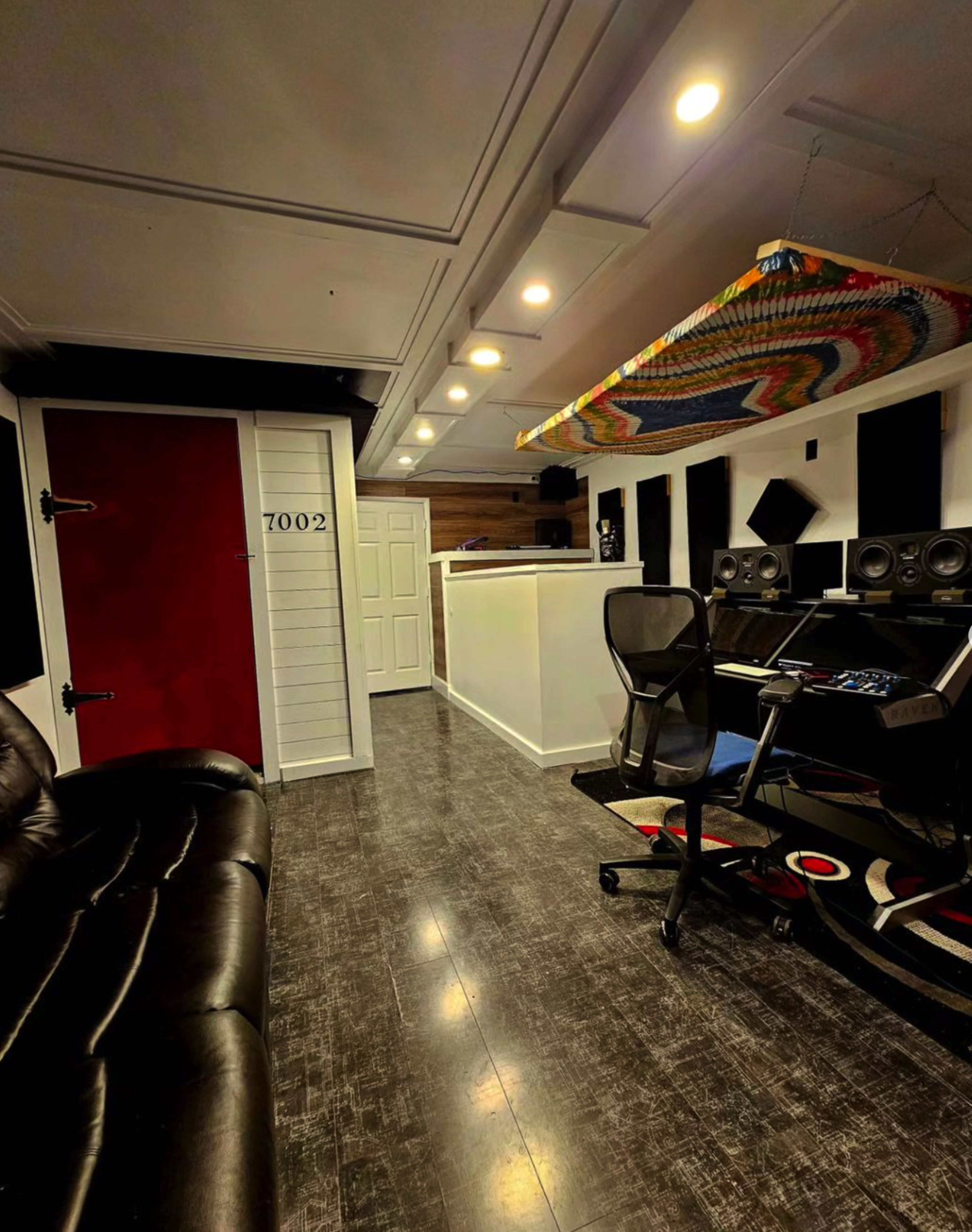 A modern recording studio with a black leather sofa, a control desk with audio equipment, and a brightly colored woven decorative piece hanging from the ceiling.