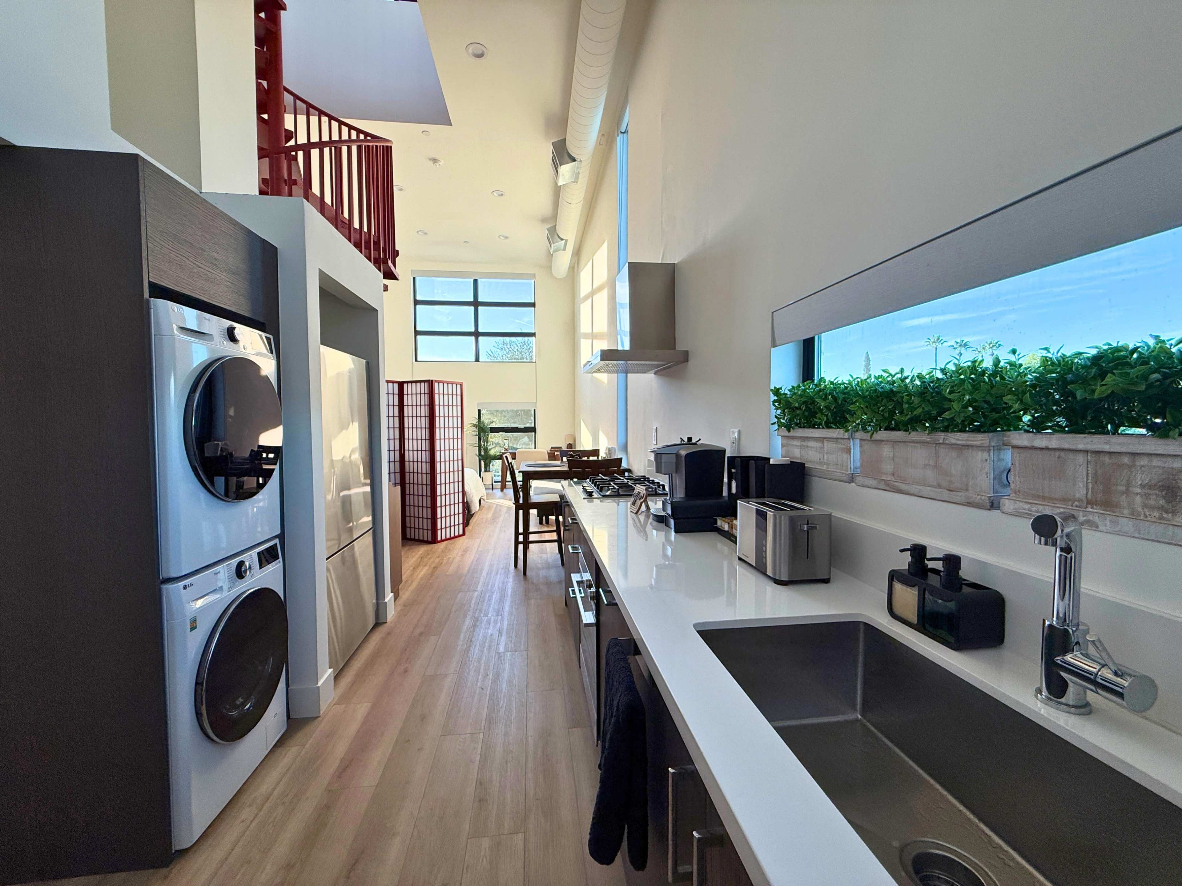 The image shows a modern kitchen with white countertops, stainless steel appliances, and a washer and dryer, leading to a dining area in a well-lit space.