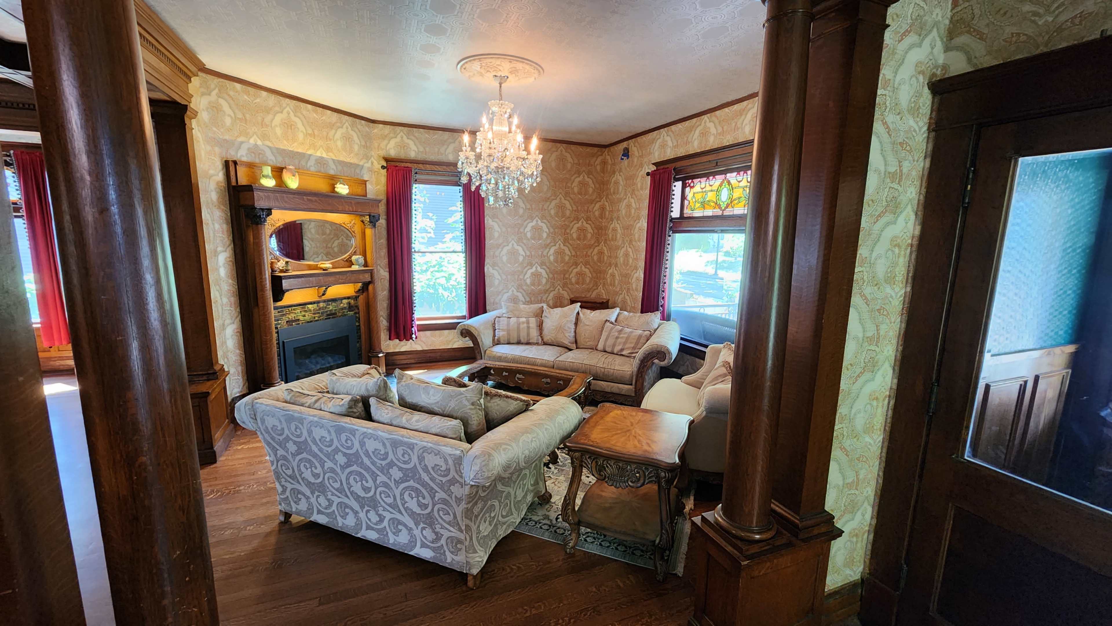 A living room with two sofas, a coffee table, and a chandelier, featuring ornate wallpaper and windows with red drapes.