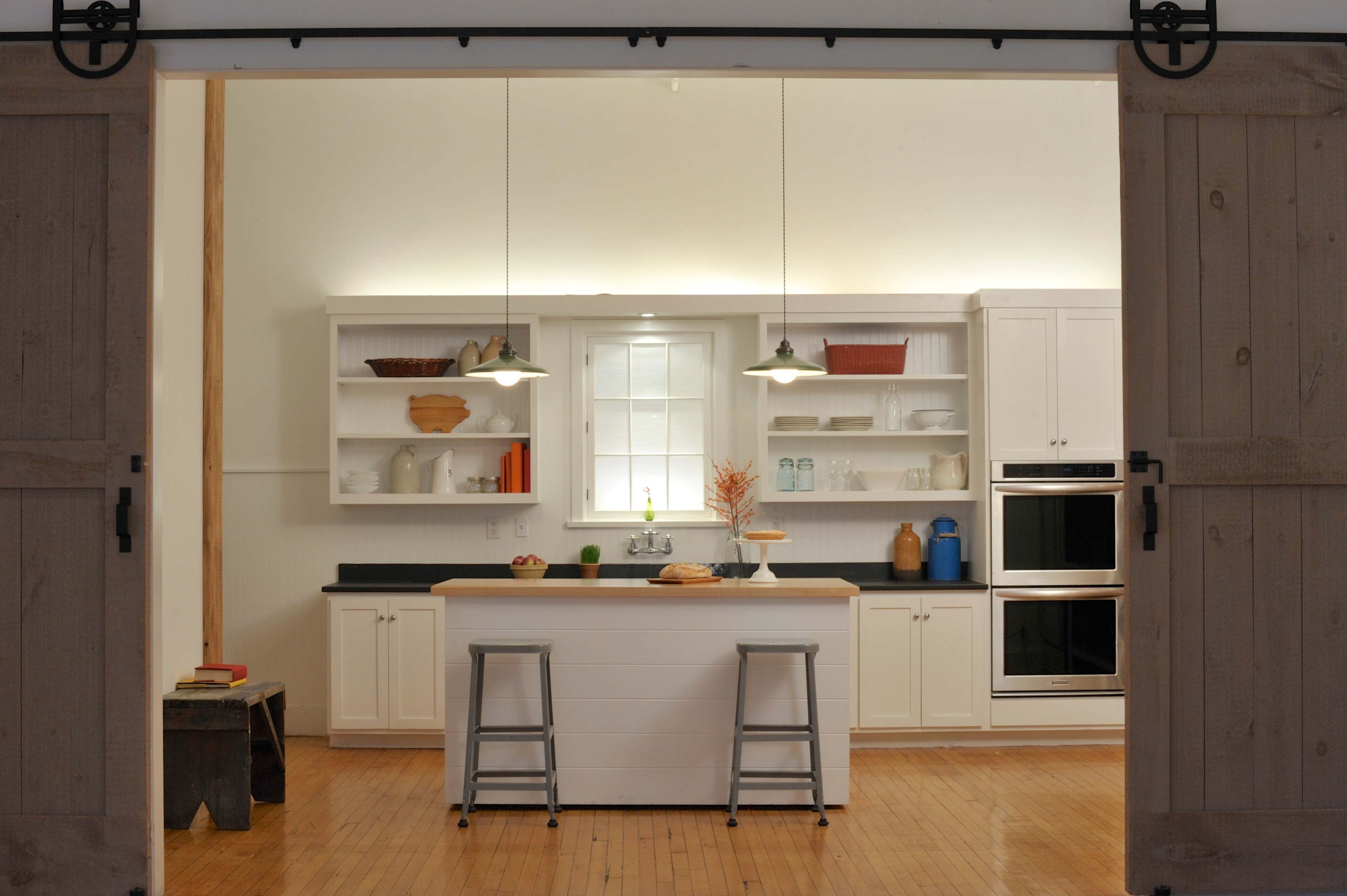 A modern kitchen features a central island with stools, white cabinetry, and open shelving against a light backdrop.