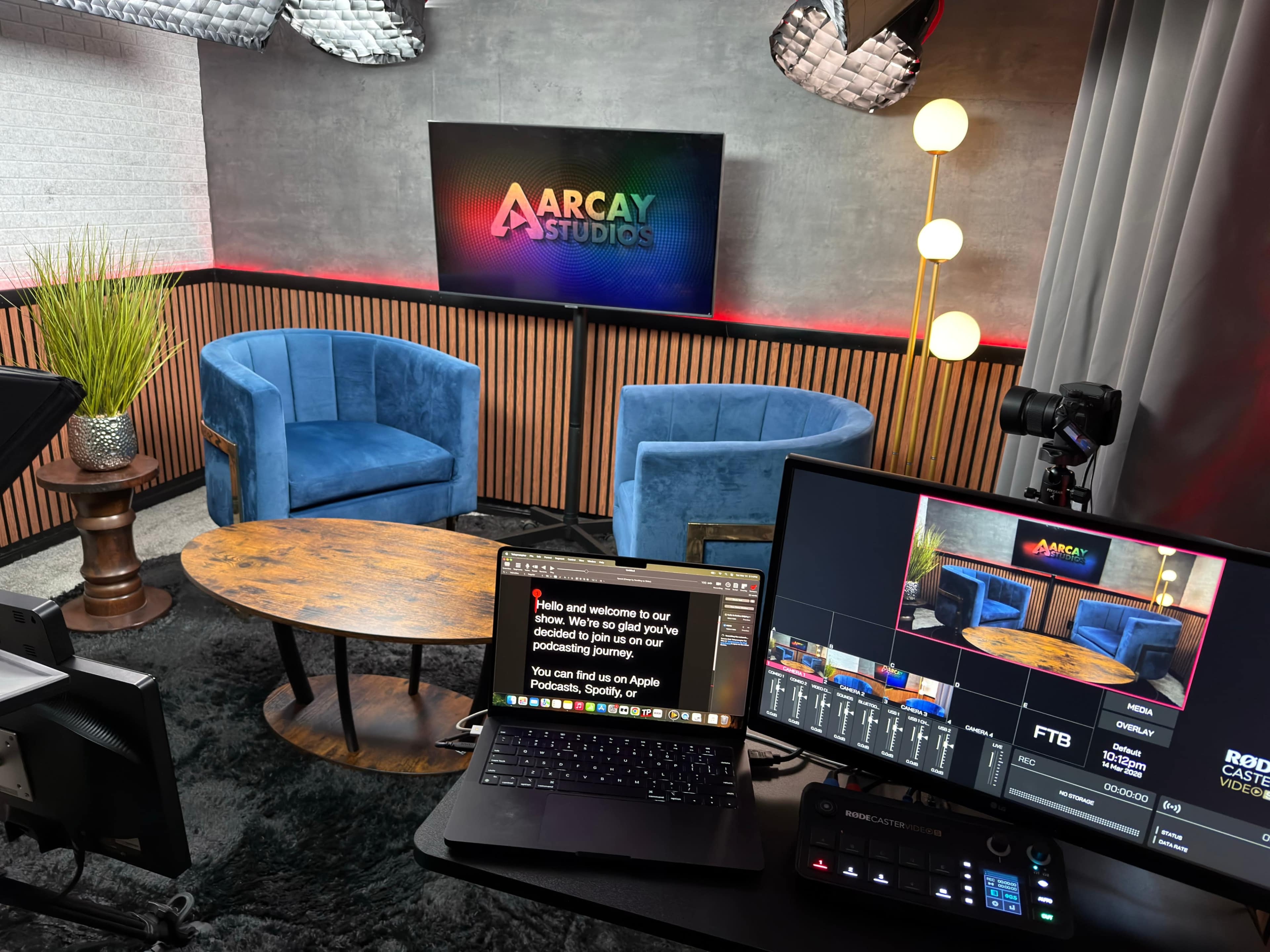 The image shows a modern studio setup with blue velvet chairs, a wooden table, a large display with the "ARCAY Studios" logo, and broadcasting equipment including a laptop and camera.