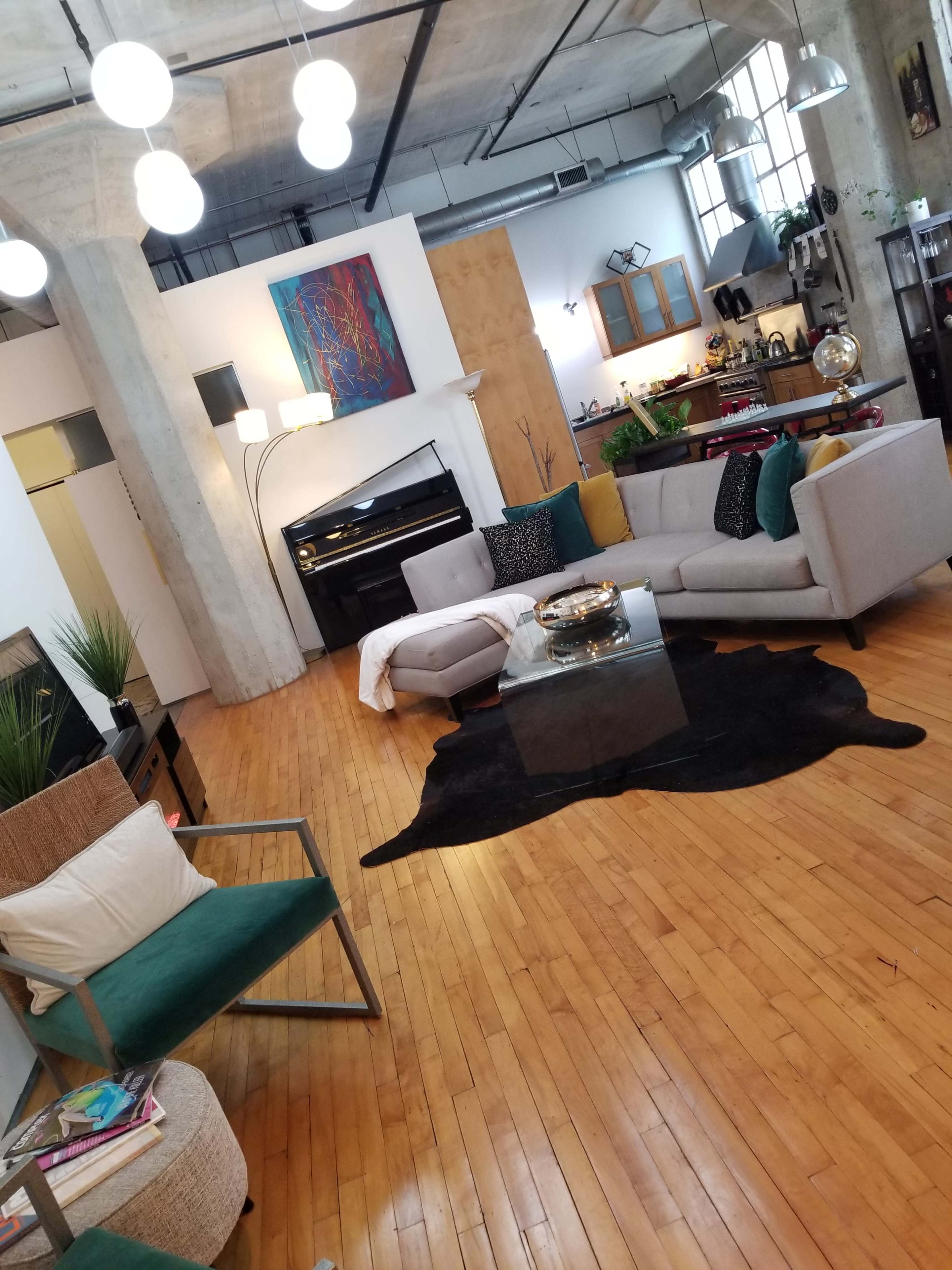 A modern living space with a large sectional sofa, a central coffee table on a black rug, and various decorative elements against a backdrop of hardwood floors and exposed concrete pillars.