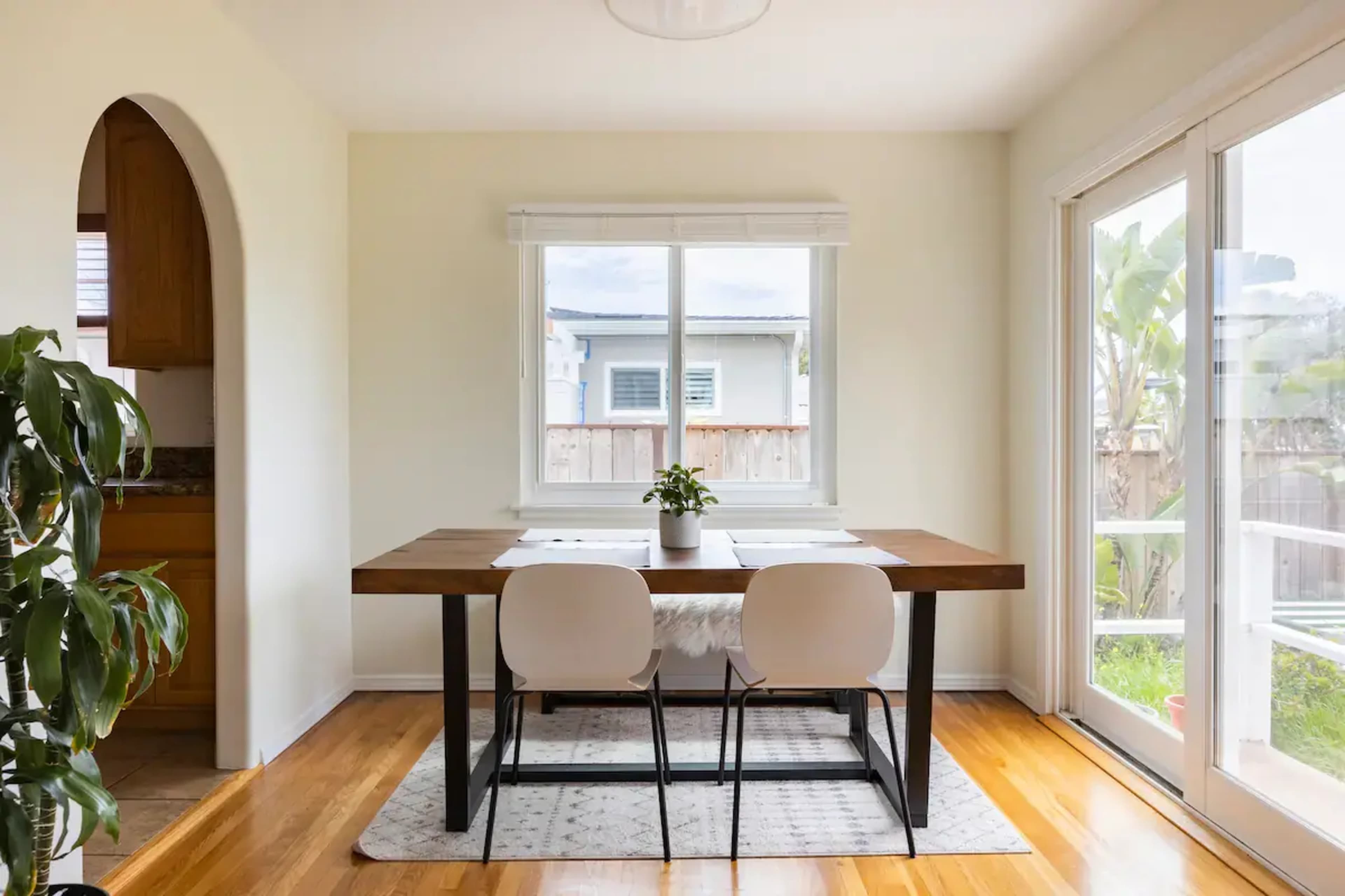 A wooden dining table is set with two white chairs, positioned in a bright room with large windows and a potted plant nearby.