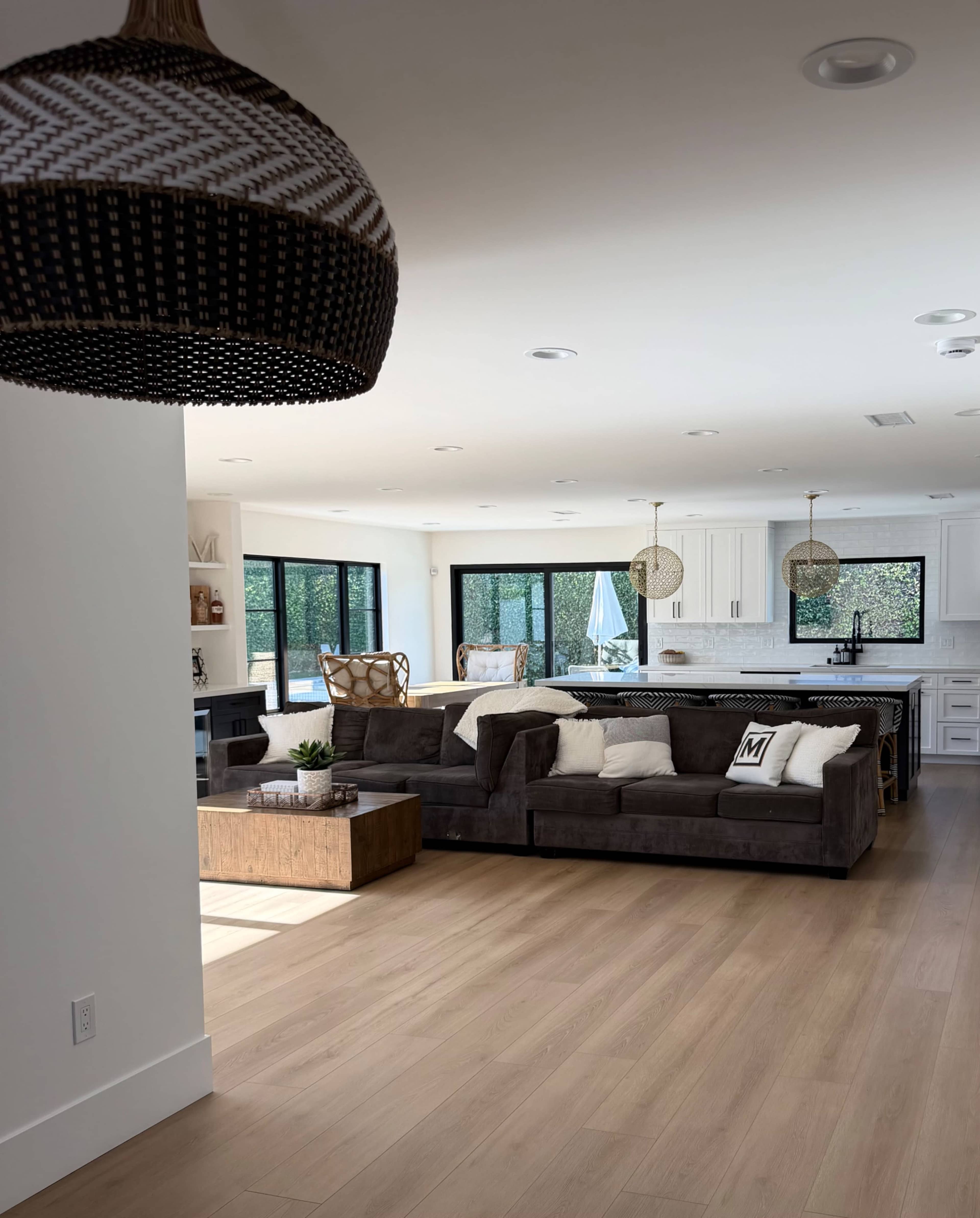 The image shows a modern, open-concept living space featuring a dark sectional sofa, a wooden coffee table, and a well-lit kitchen in the background.