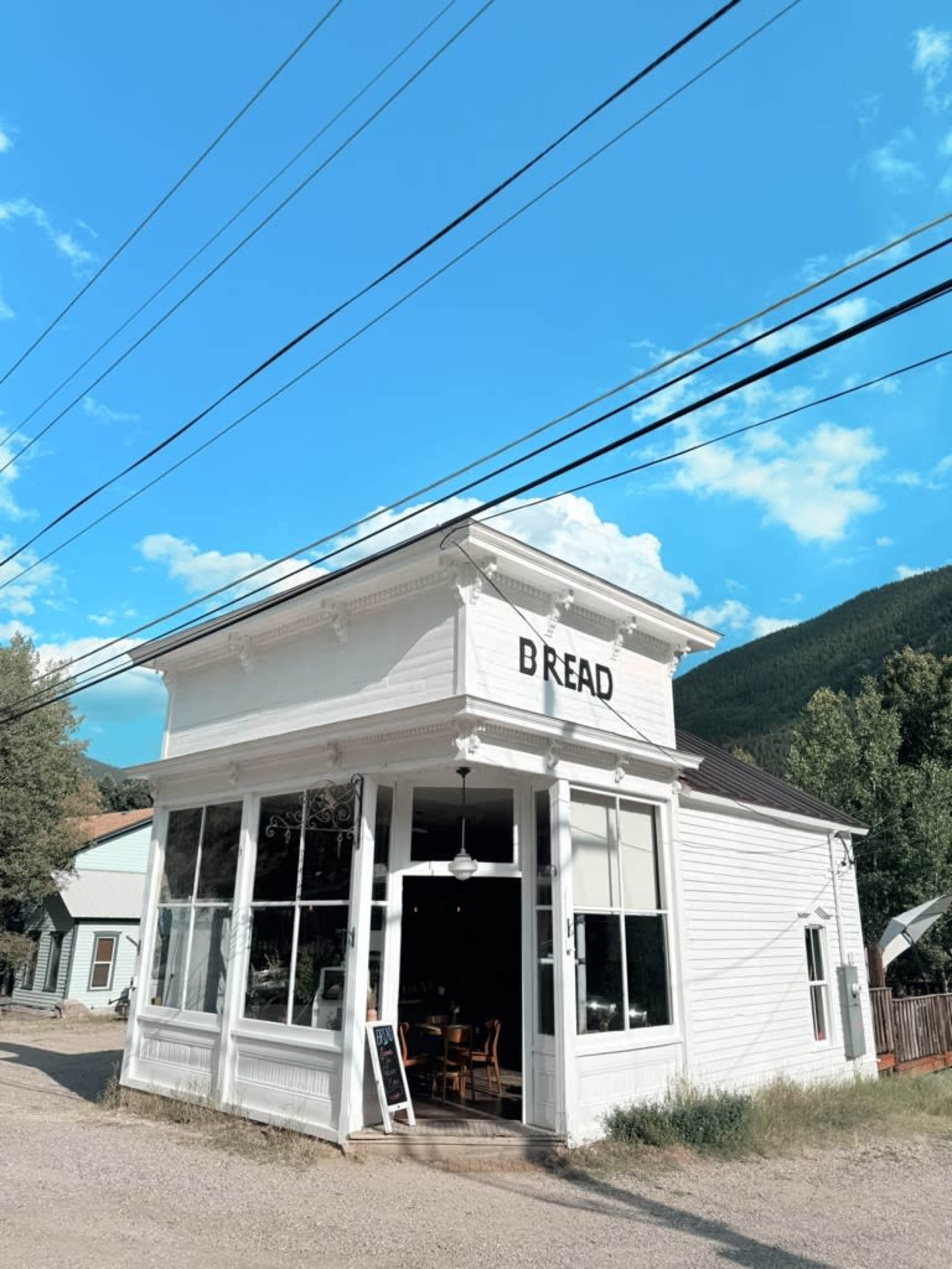 The image shows a white, corner storefront labeled "BREAD," surrounded by trees and a blue sky with clouds.