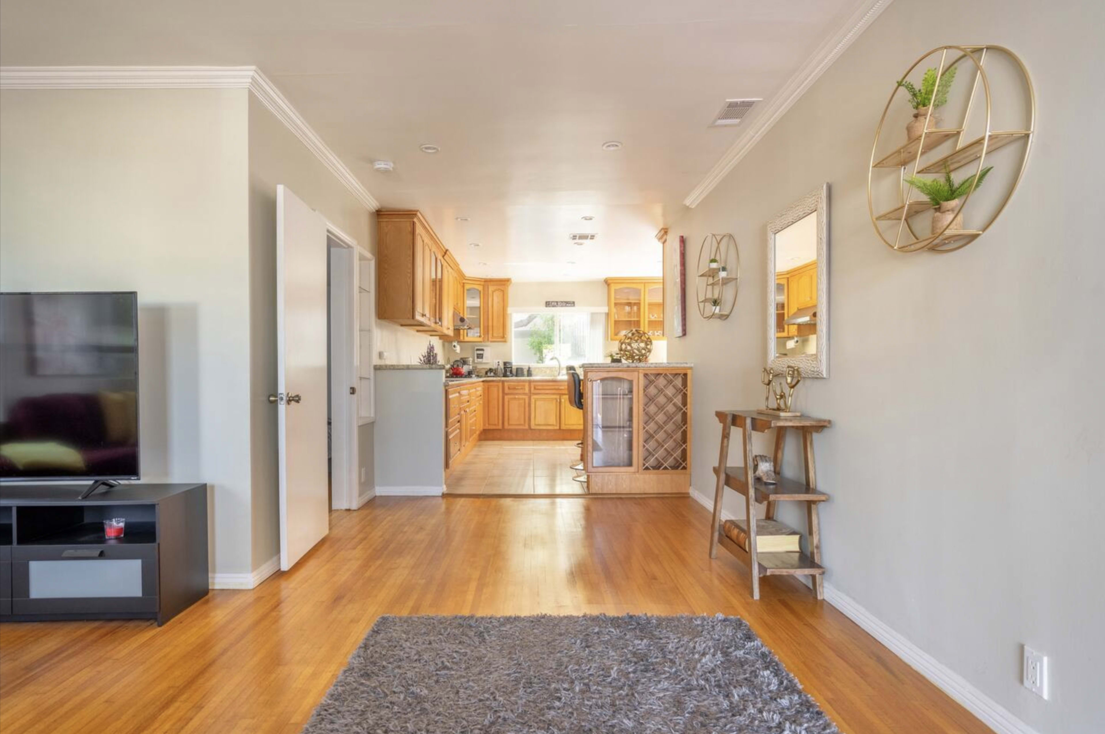The image shows an open-concept living area with hardwood floors leading to a kitchen in the background, featuring wooden cabinets and a small dining space.