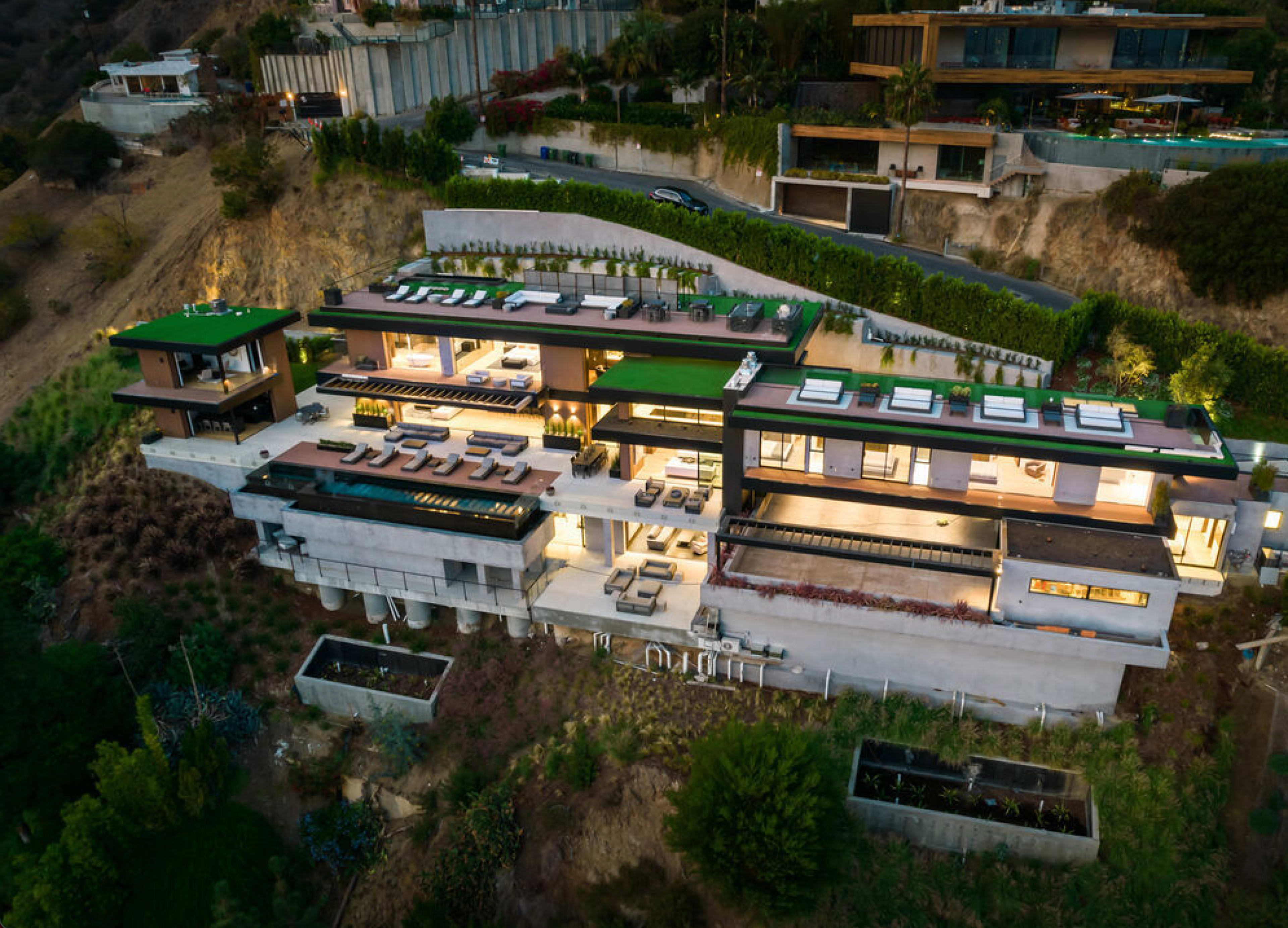 The image depicts a modern, multi-level luxury home situated on a hillside, featuring a green rooftop and expansive outdoor seating areas.
