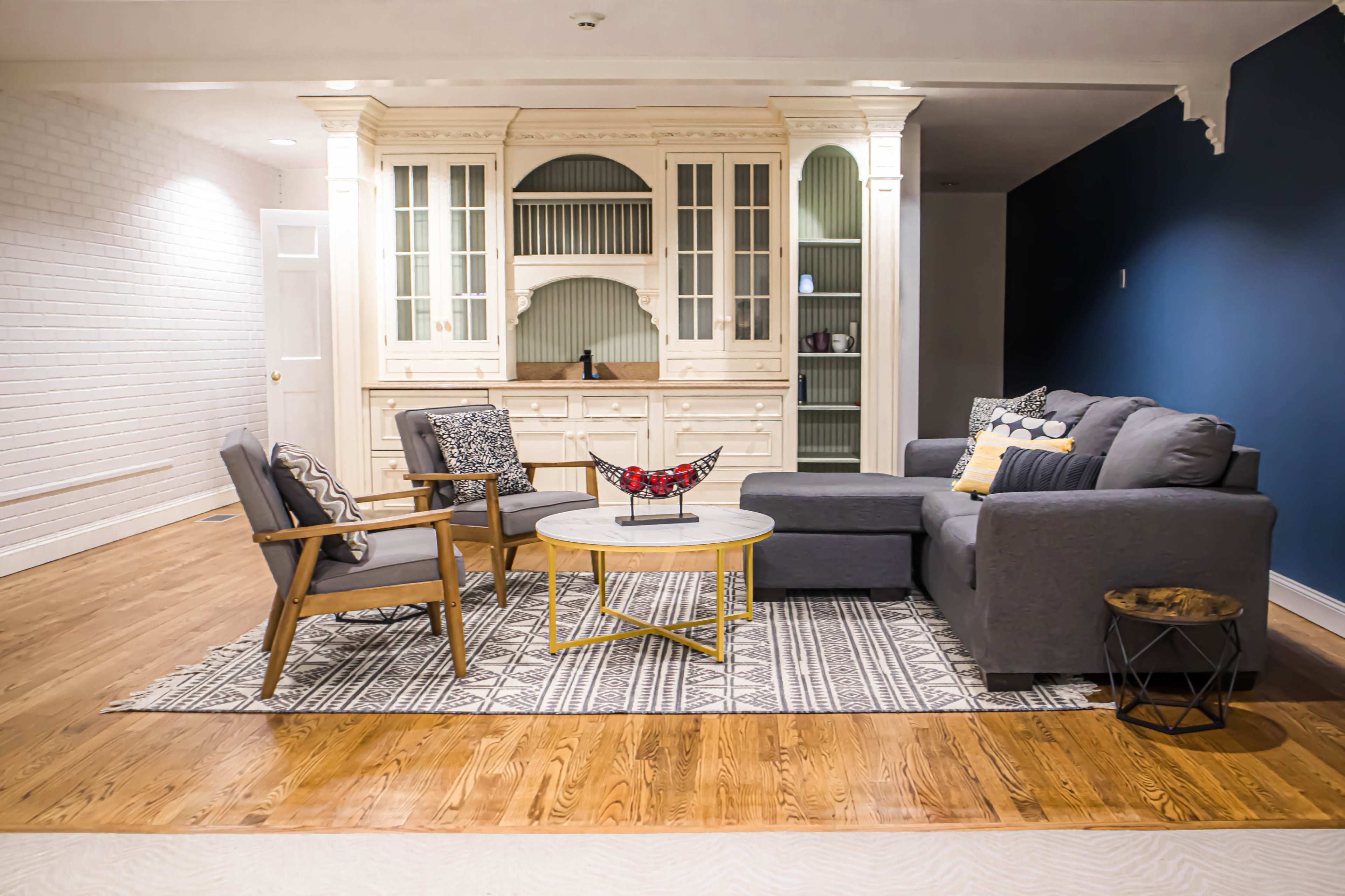 A modern living room with a gray sofa, two wooden chairs, a round coffee table, and a light-colored cabinet against a dark blue wall.