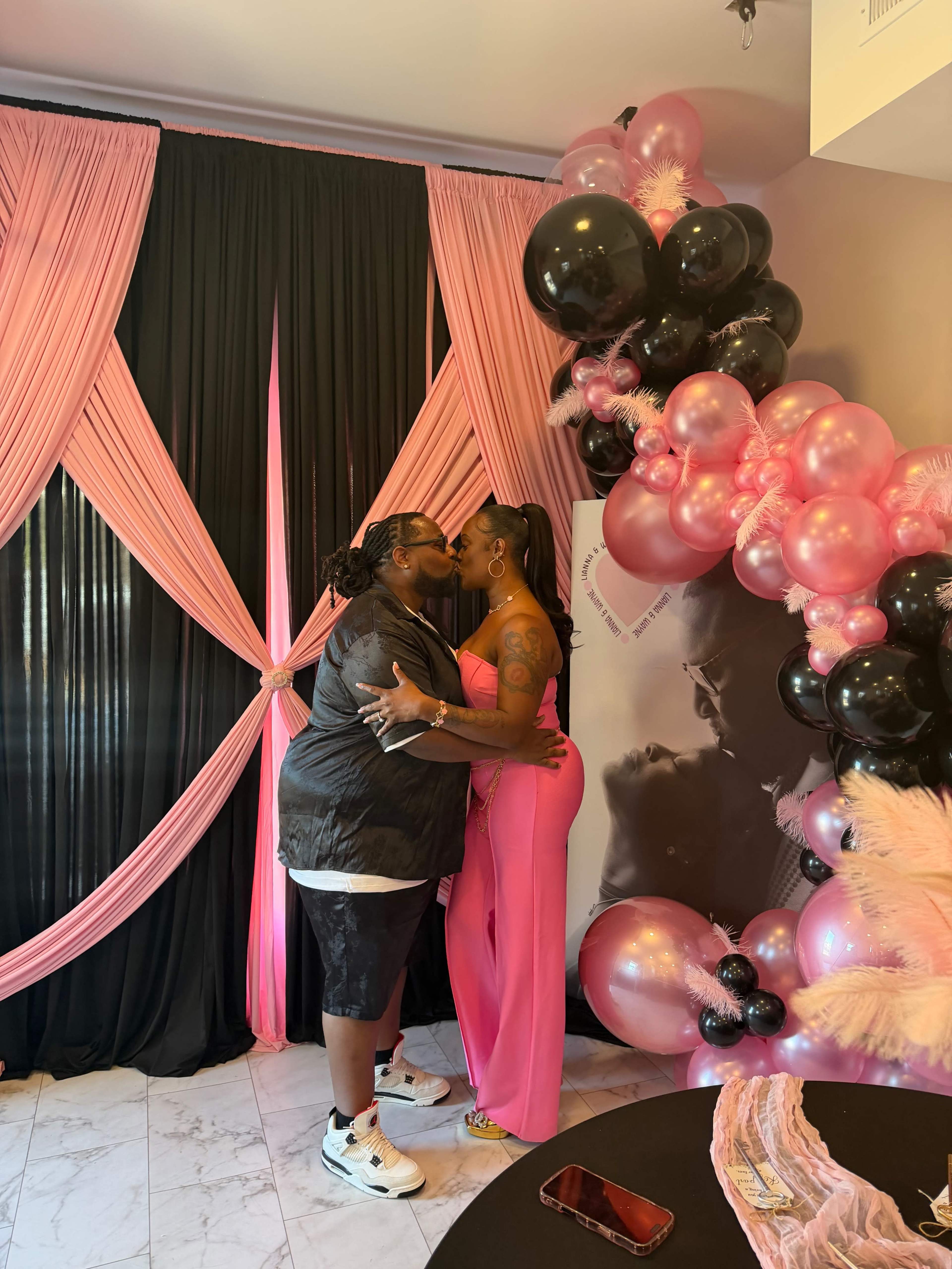A couple shares a kiss in a decorated space featuring pink and black drapes and balloons.