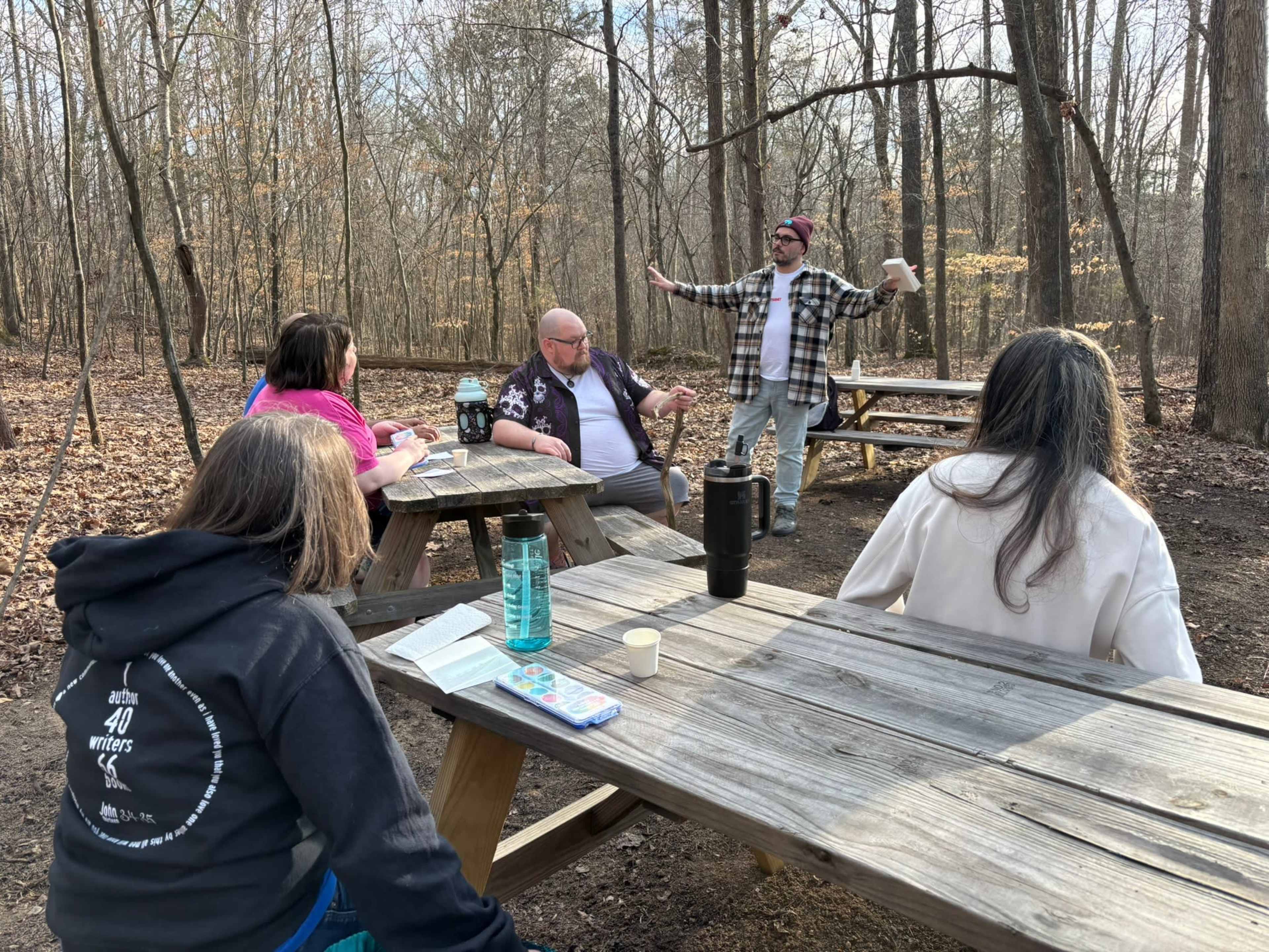 A group of people sits at wooden picnic tables in a wooded area while one person stands and gestures, speaking to the group.