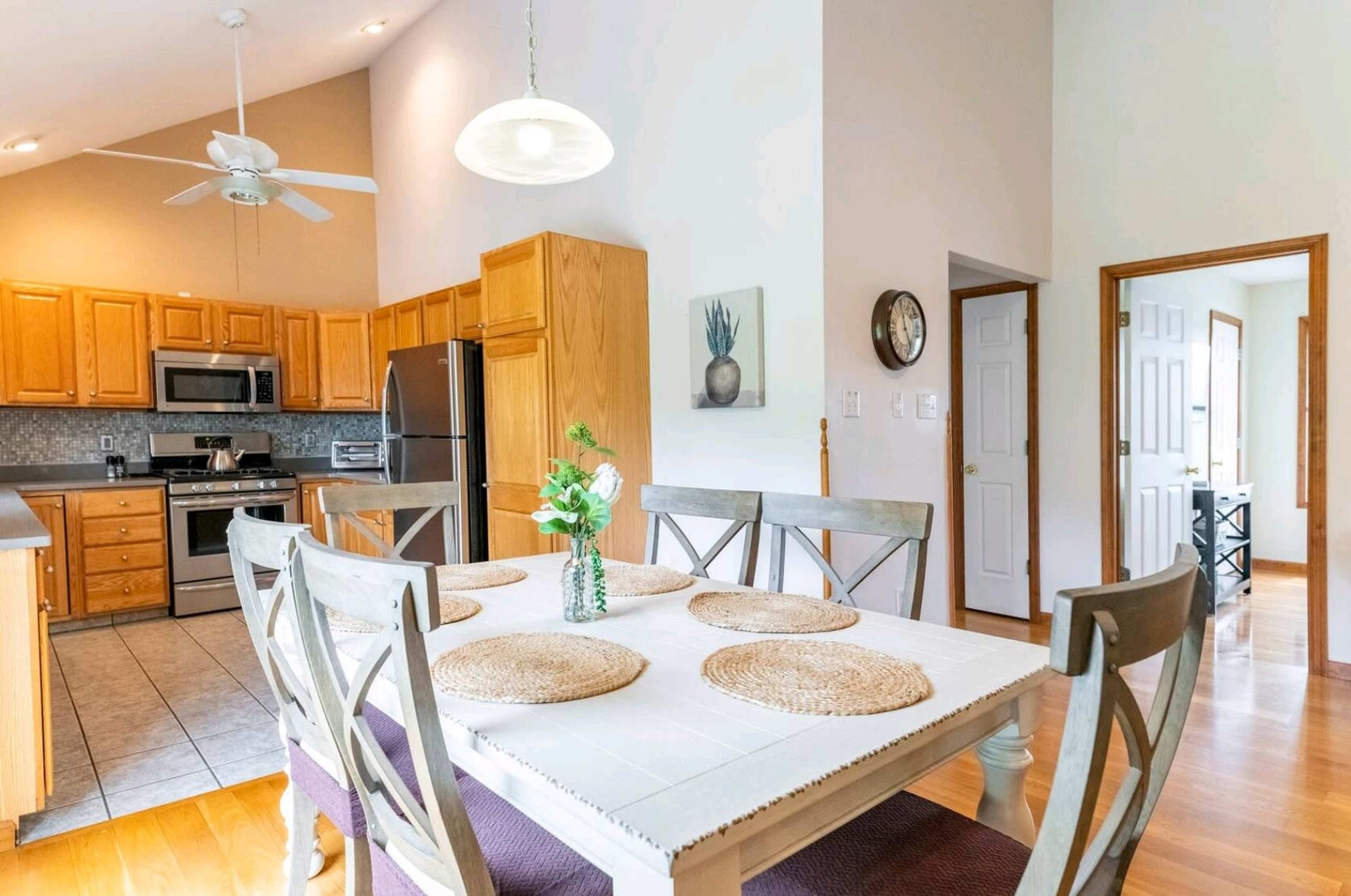 A dining area features a wooden table set with placemats, surrounded by chairs, and is adjacent to a kitchen with light wood cabinets and stainless steel appliances.