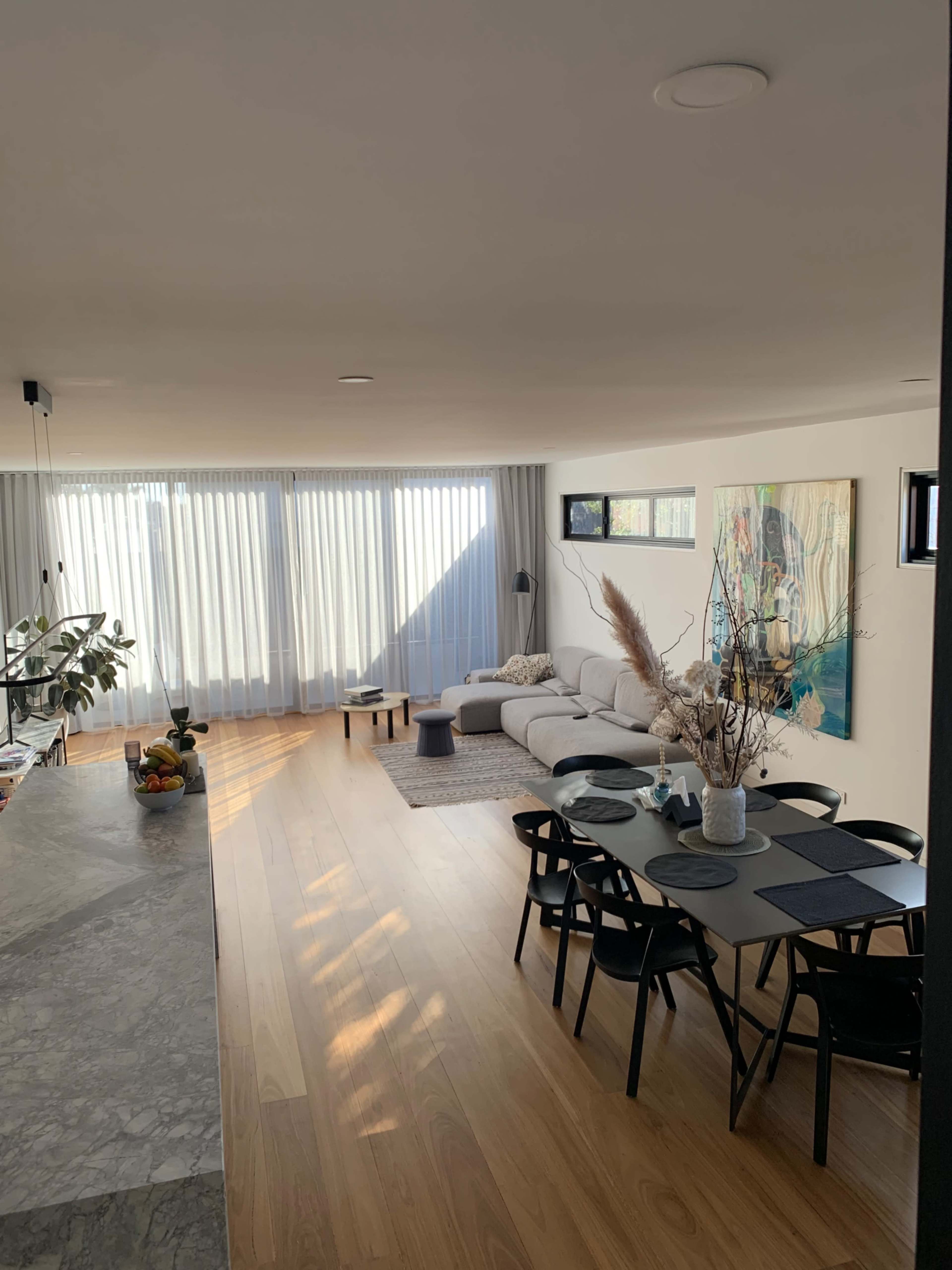 A modern living room with a light-colored sofa, a dining table with black chairs, and large windows covered by sheer curtains that allow natural light into the space.