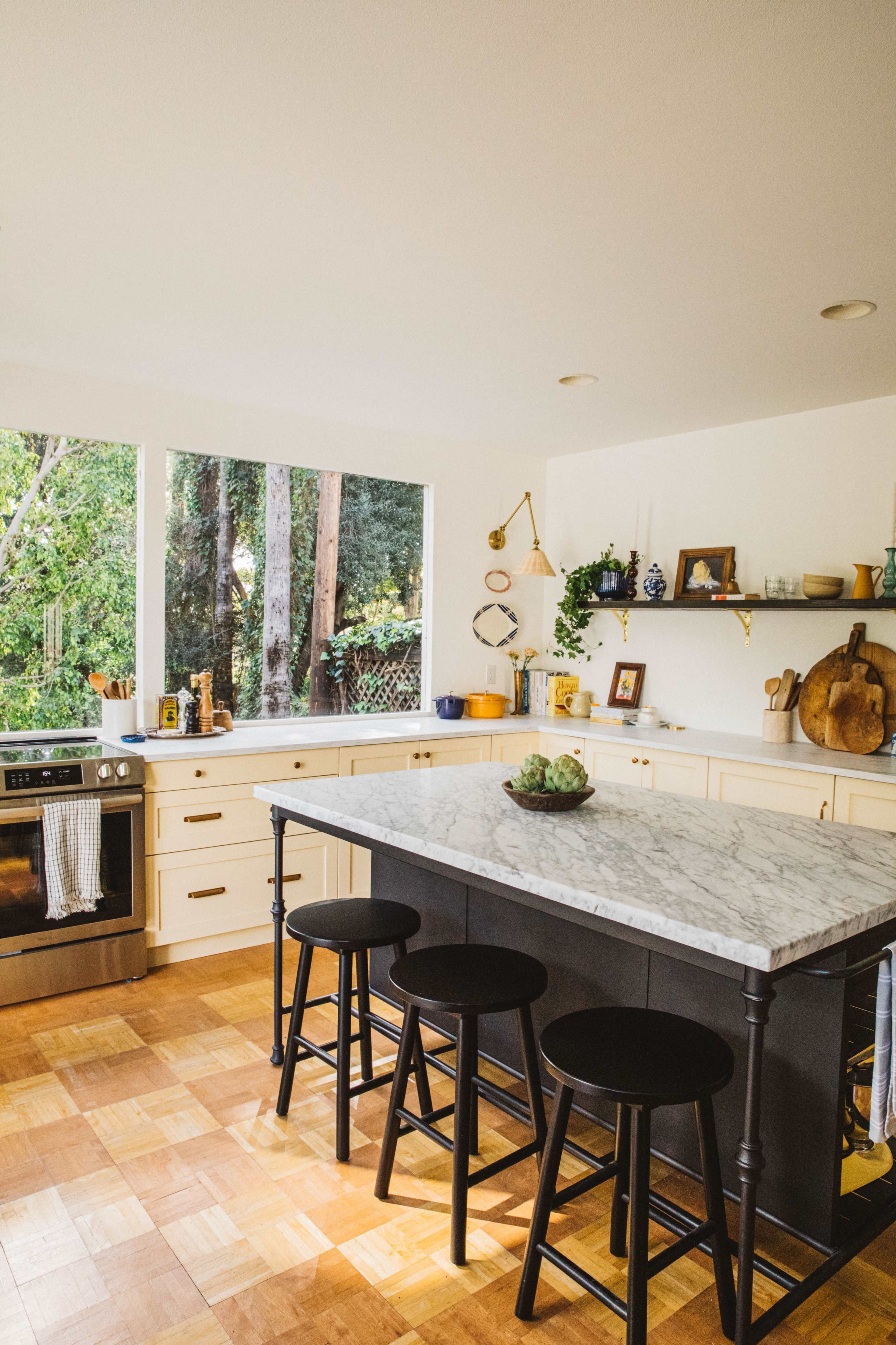 The modern kitchen features a marble-topped island with three black stools, surrounded by a backdrop of large windows revealing greenery outside.