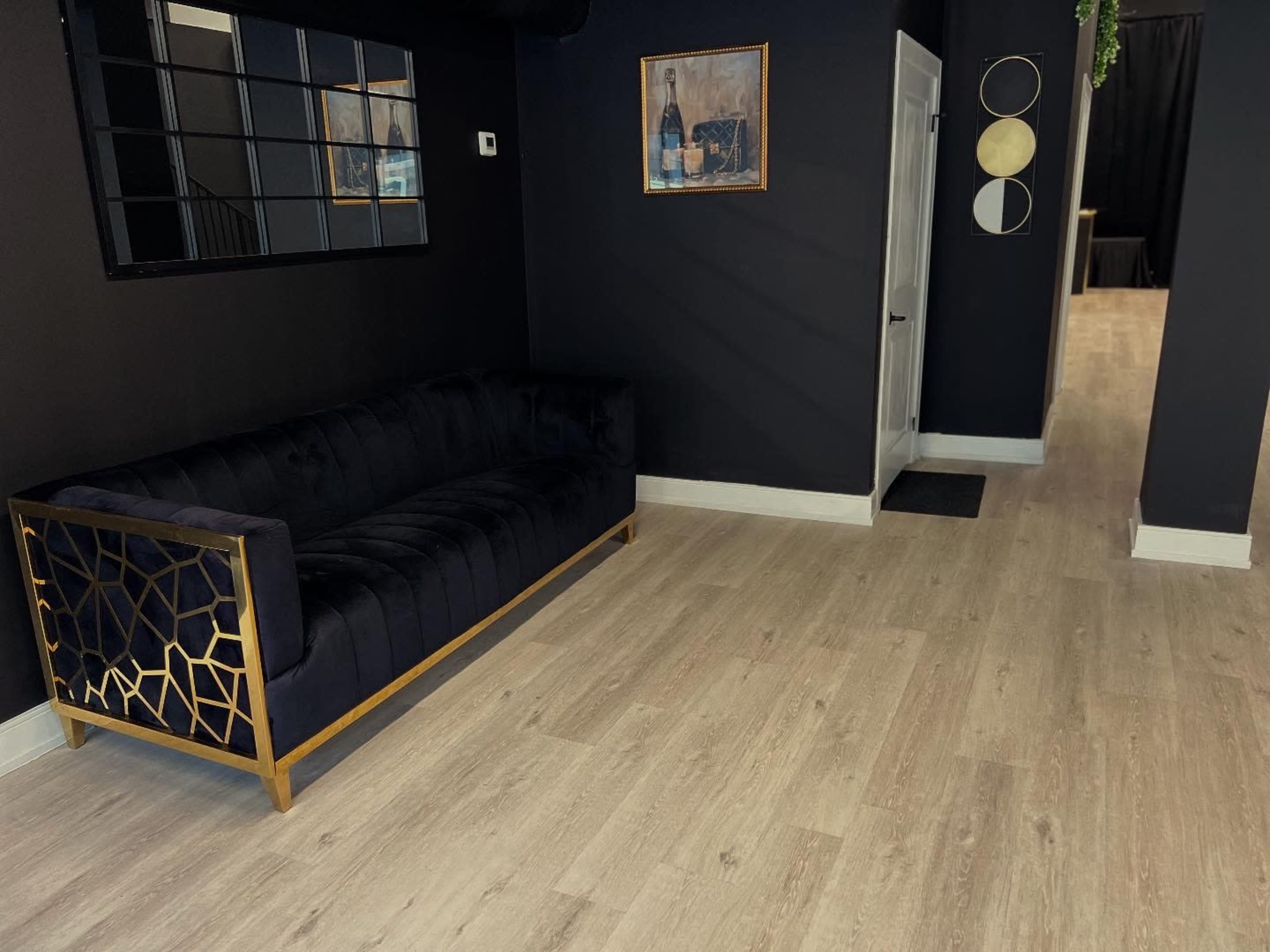 The image shows a black-walled interior featuring a modern gold-accented couch and wooden flooring, with a mirror and artwork on the wall.