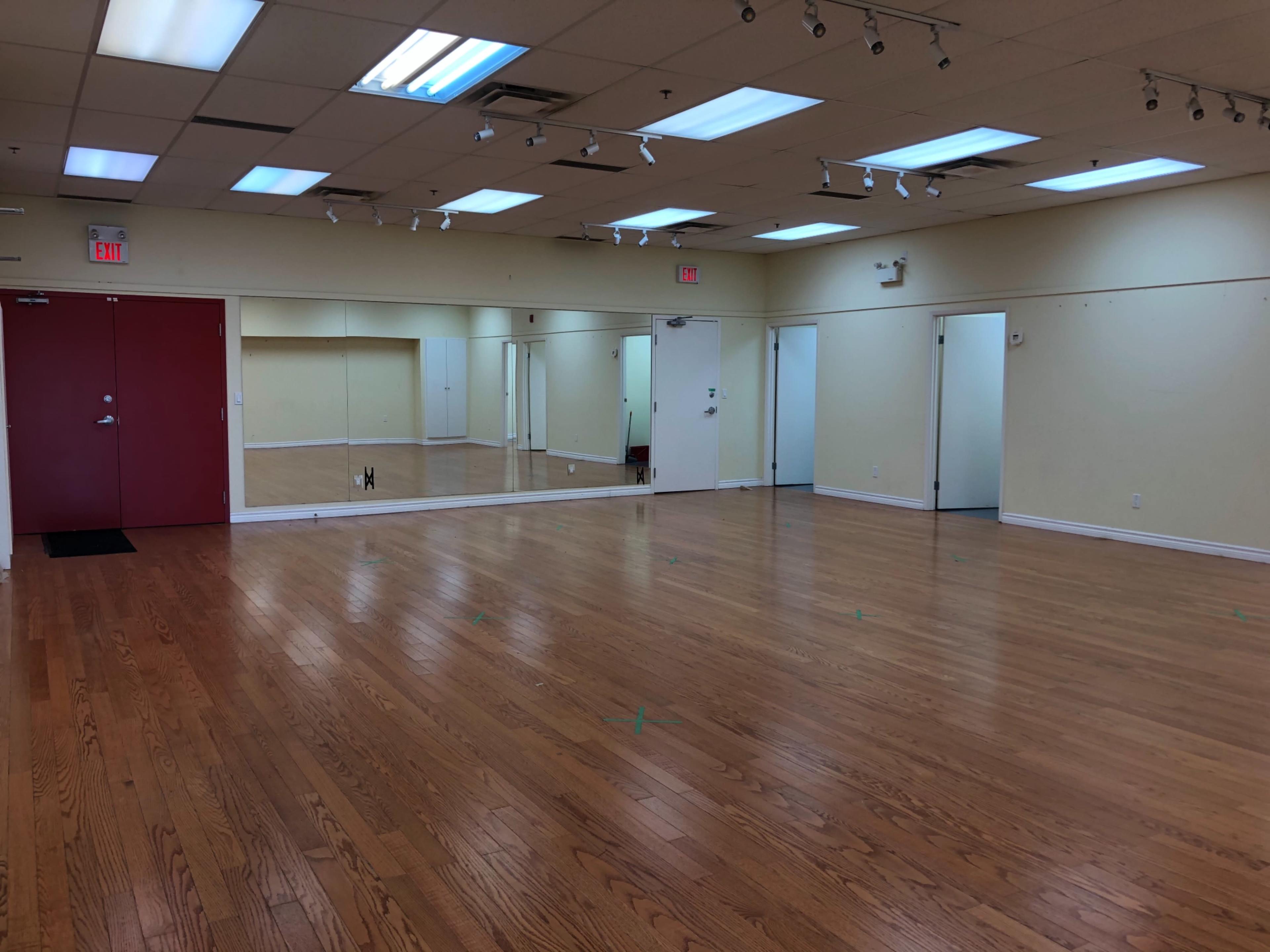 The image shows a spacious, empty room with hardwood floors, bright overhead lights, and several mirrors on one wall.