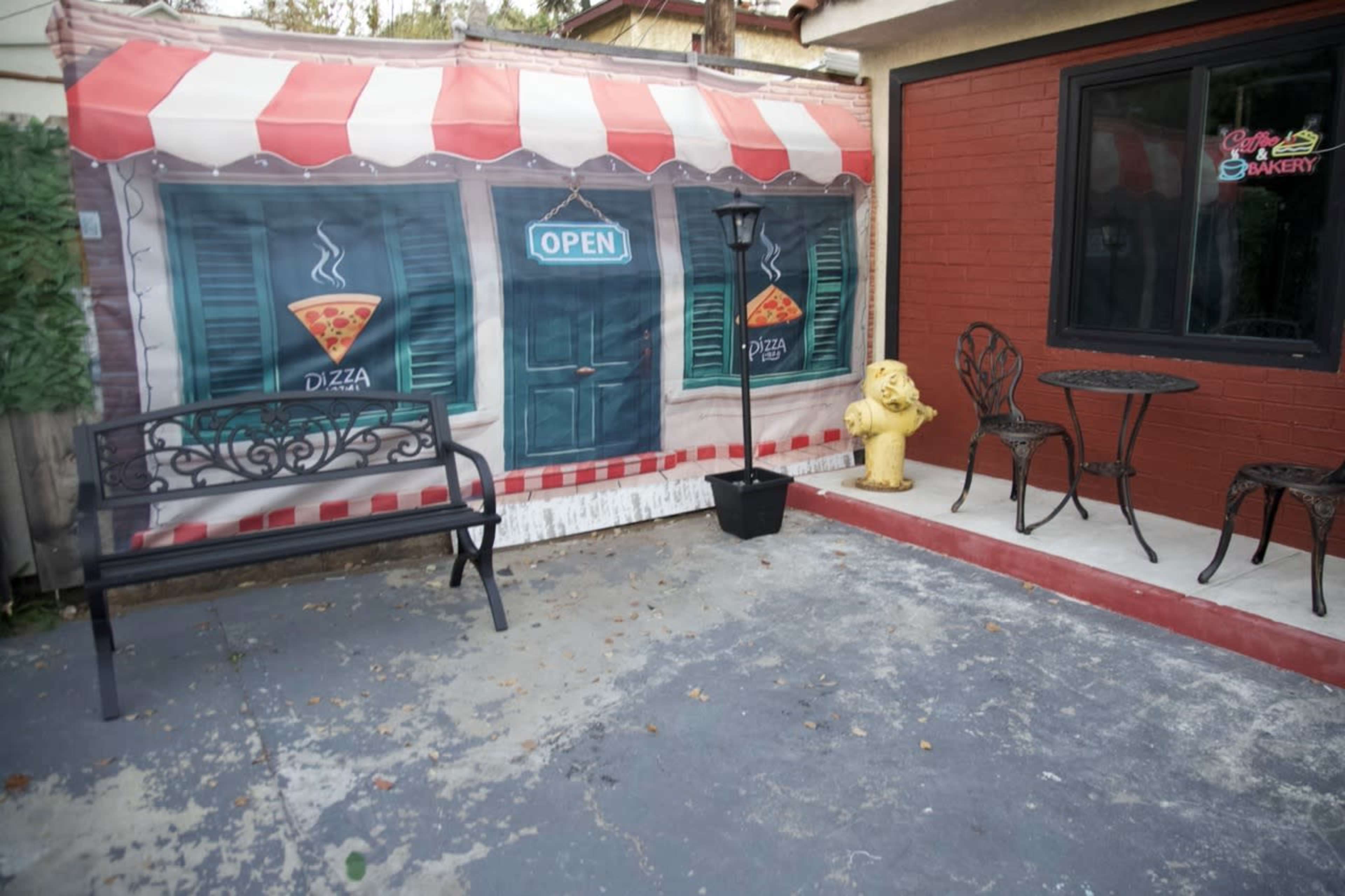 The scene features a patio area with a mural of a pizza shop, a bench, a table with chairs, and a yellow fire hydrant nearby.