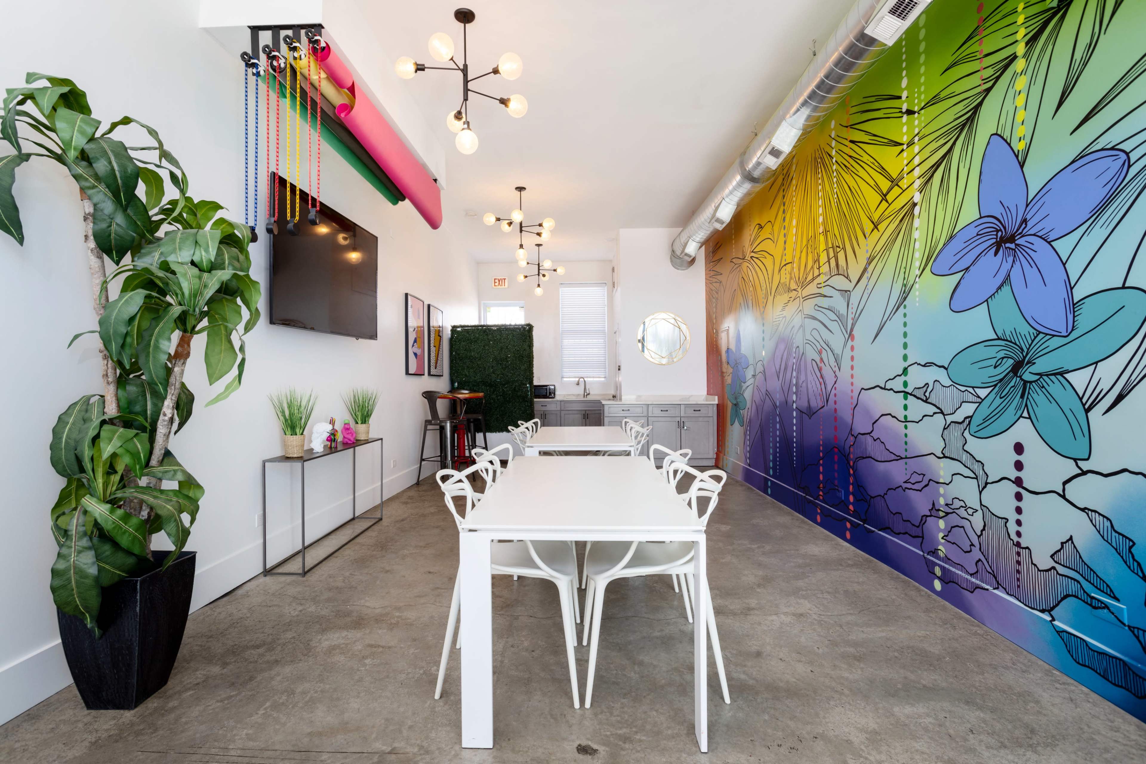 A brightly lit room featuring a long white table with chairs, a colorful mural on one wall, and a green plant in the corner.