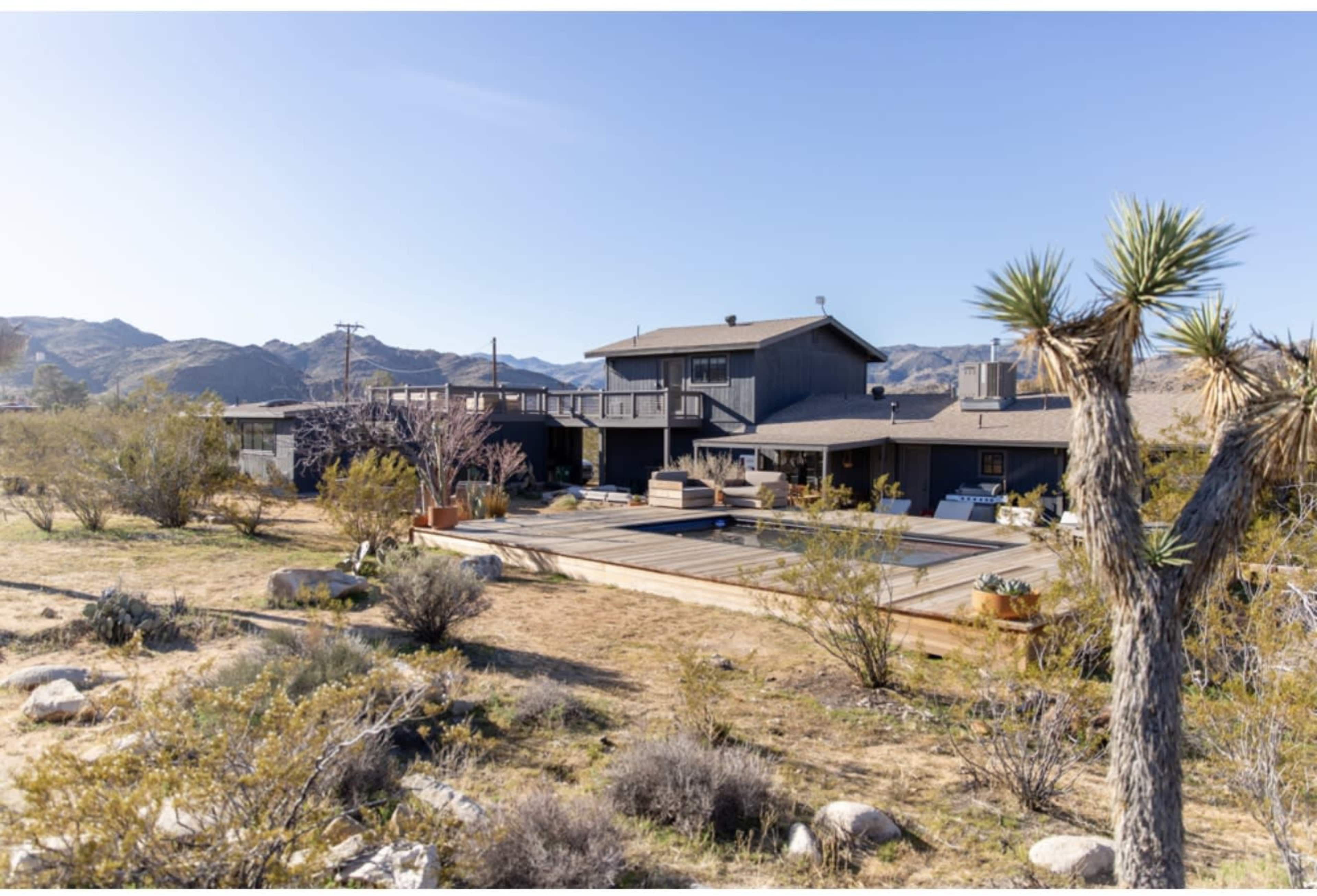 A two-story house with a pool is situated in a desert landscape surrounded by shrubs and mountains in the background.
