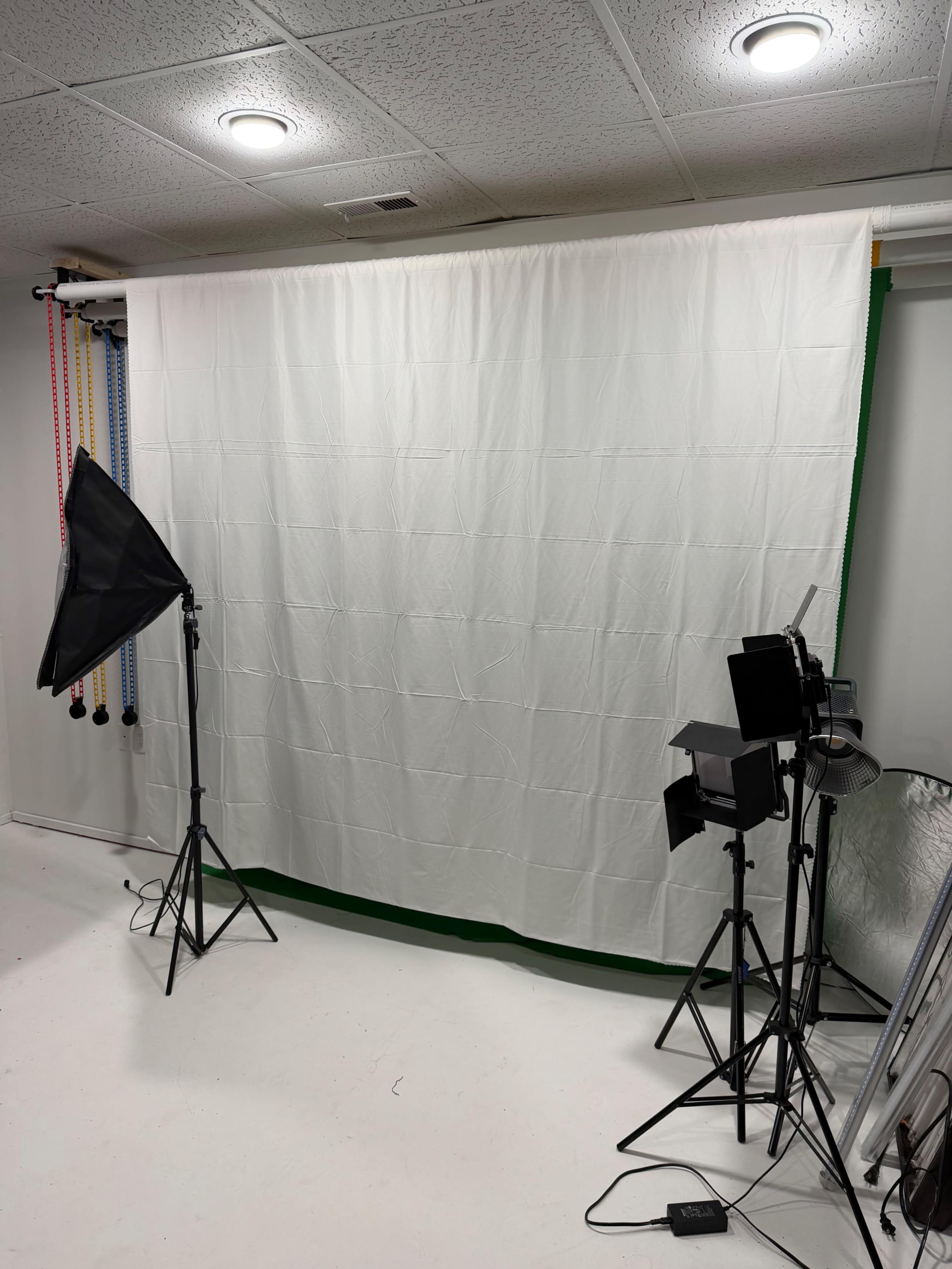 The image shows a photography studio set up with a white backdrop, lighting equipment on tripods, and other gear positioned for a photoshoot.