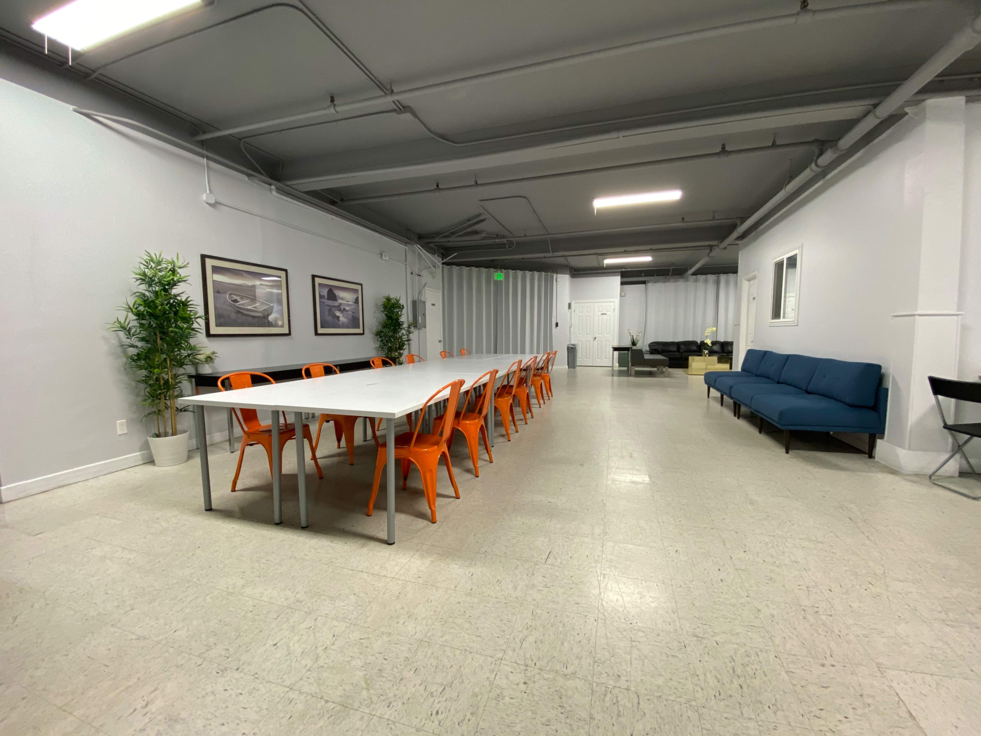A spacious room with a long table surrounded by orange chairs, a blue couch, and decorative plants against the walls.