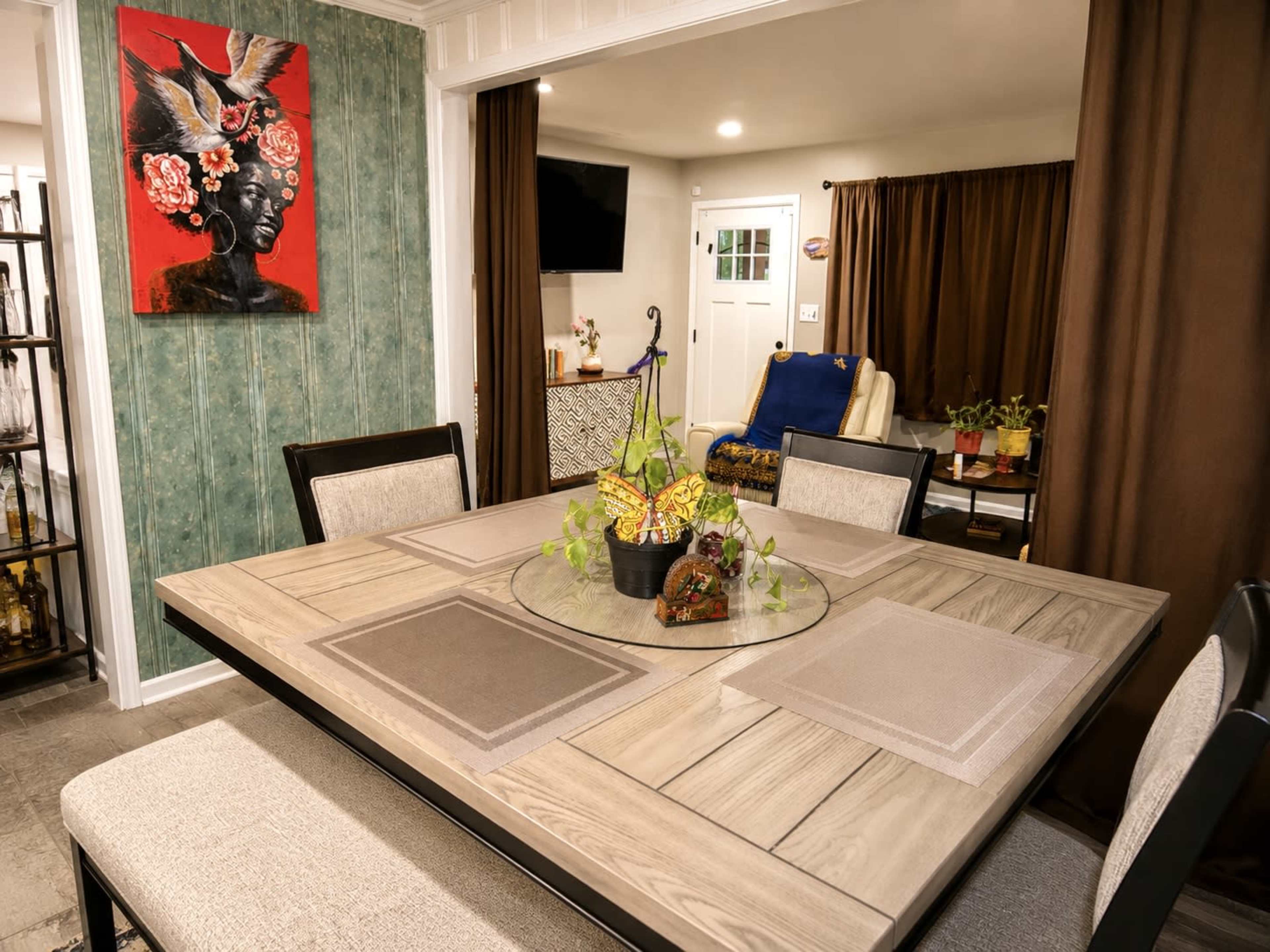 The image shows a dining area with a wooden table, a decorative centerpiece, and a vibrant wall art piece, adjacent to a living room with potted plants and a television.