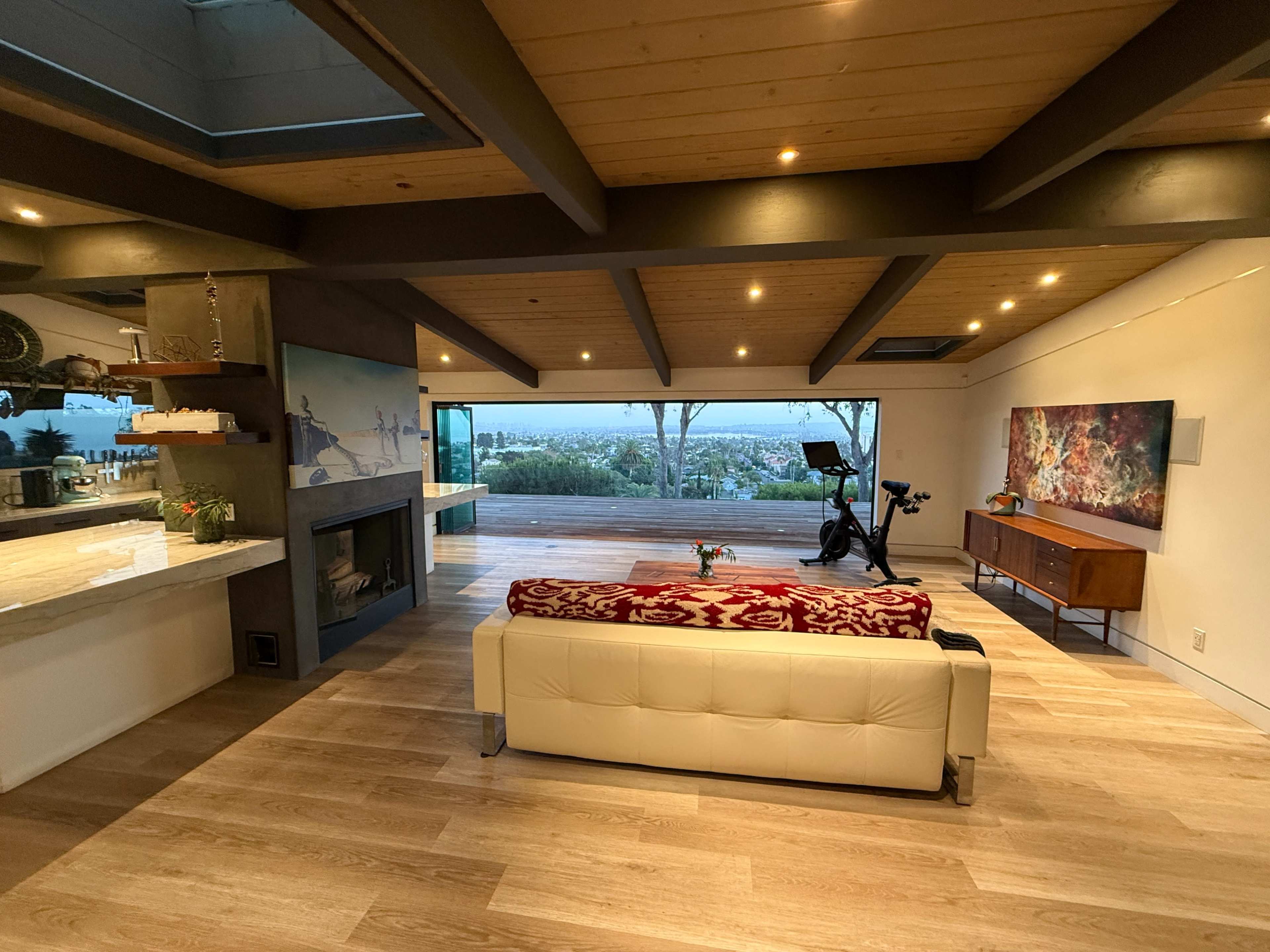 The image depicts a modern living room with a white couch, wooden ceiling beams, a fireplace, and large windows offering a view of the outside.