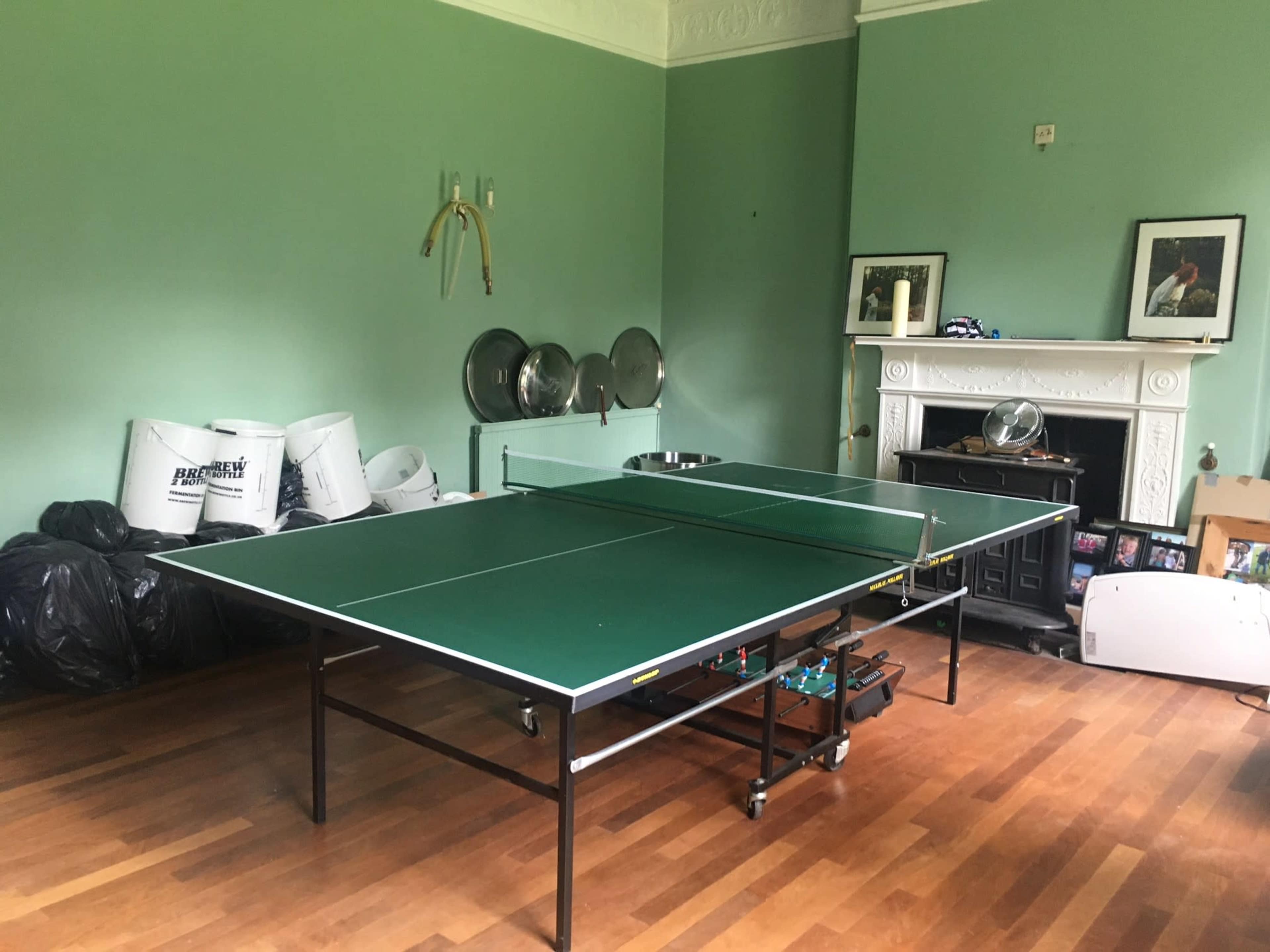 A green ping pong table set up in a room with hardwood floors and various items, including bags and framed pictures, in the background.
