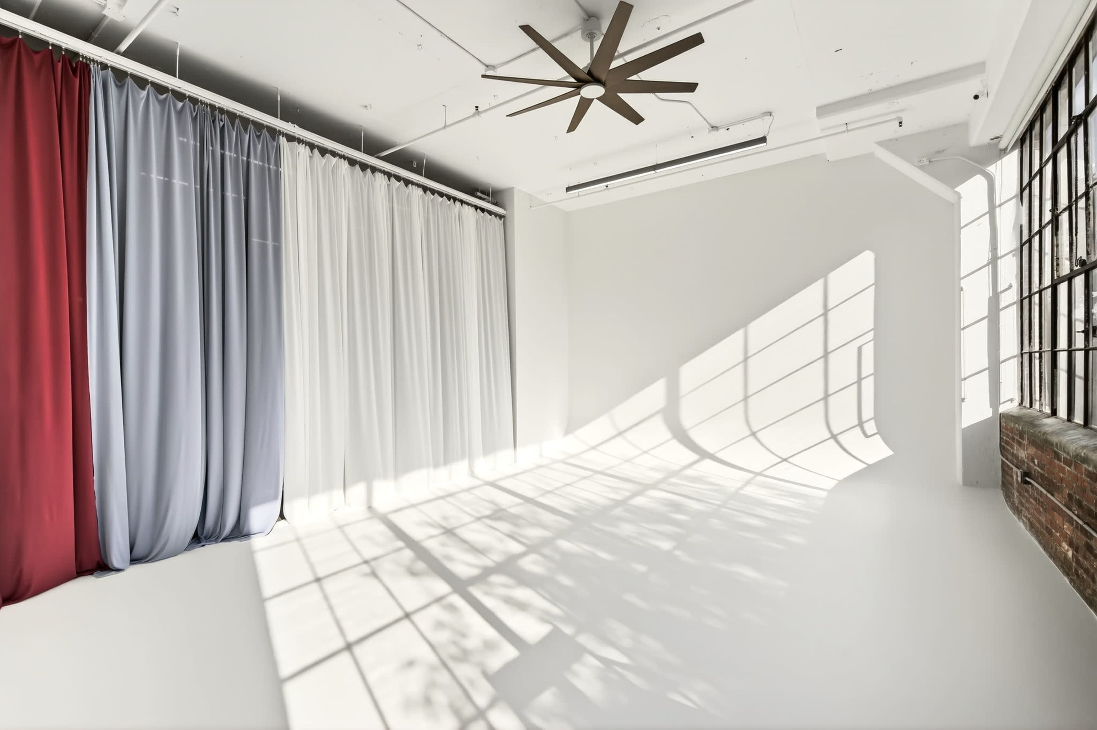 The image shows an interior space with a large window casting shadows on a white wall, complemented by curtains in red and gray.