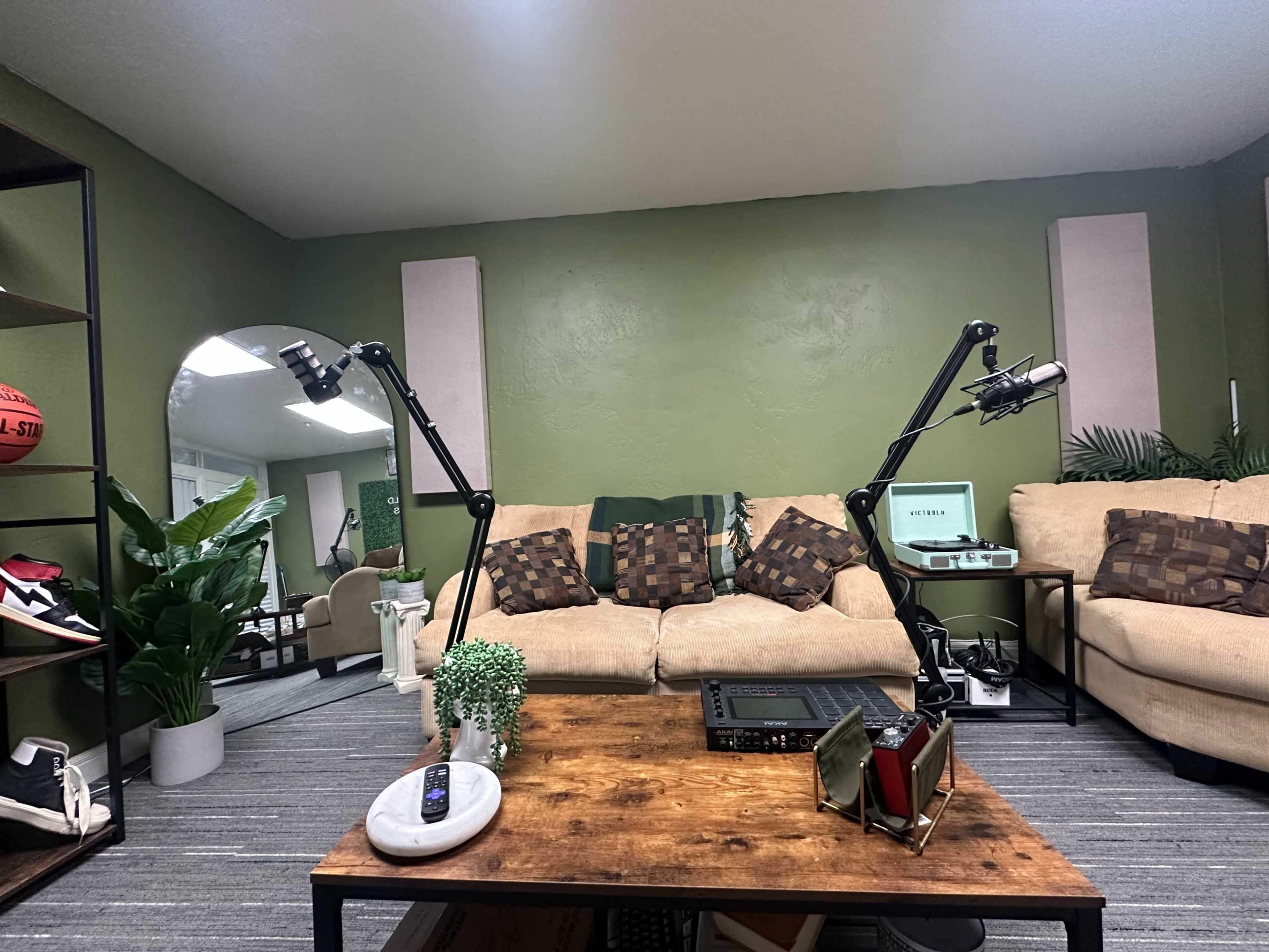 The living room features a beige couch with patterned pillows, a wooden coffee table, and two adjustable microphones on stands, with plants and sports equipment in the background.