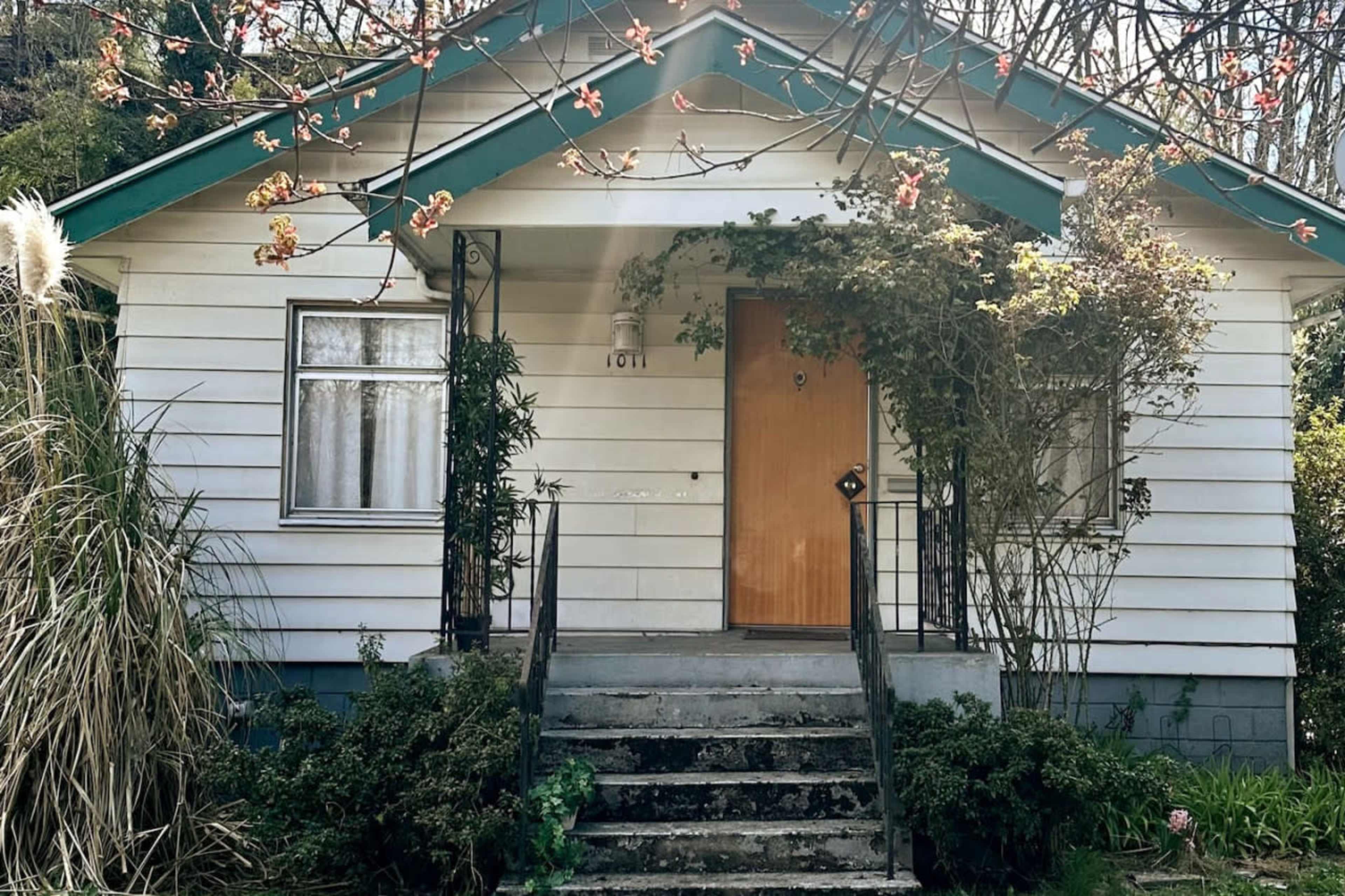 The image shows a small, single-story house with a front porch and a staircase leading up to the entrance door, surrounded by greenery and trees.