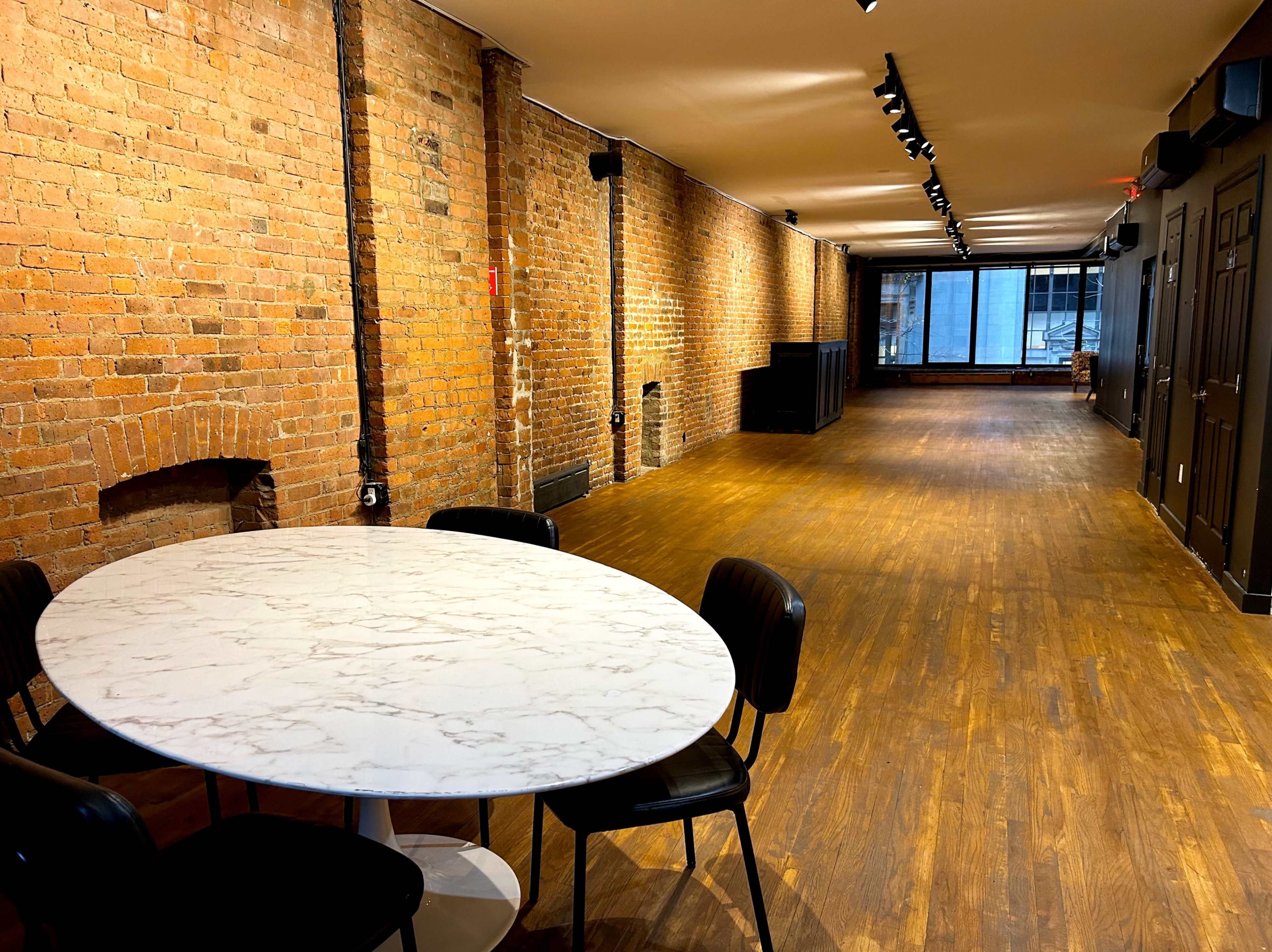 The image shows an empty room with hardwood flooring, exposed brick walls, and a round marble table with black chairs on one side.