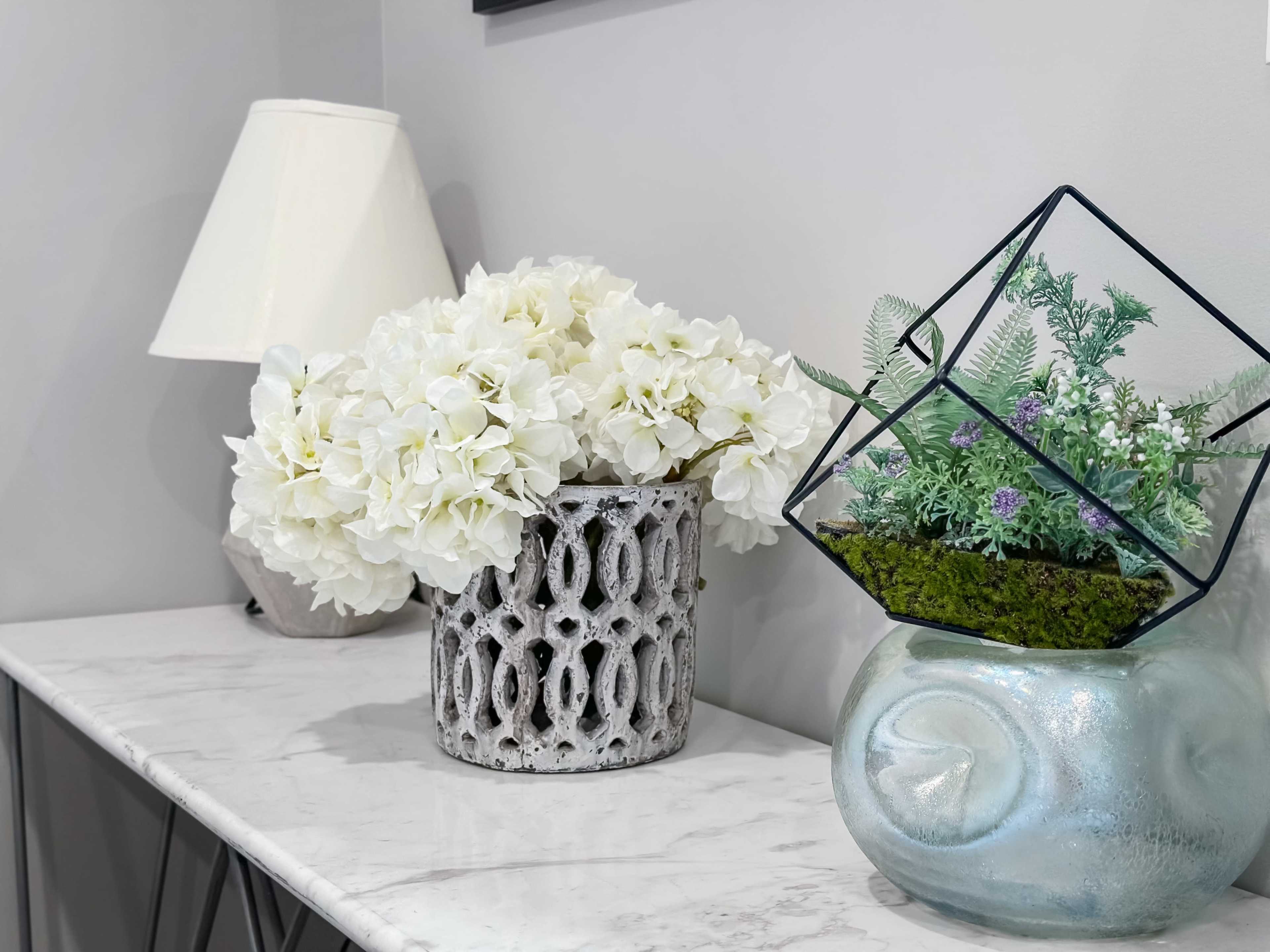 A marble table displays a decorative lamp, a textured vase of white flowers, and a geometric glass container with green plants.