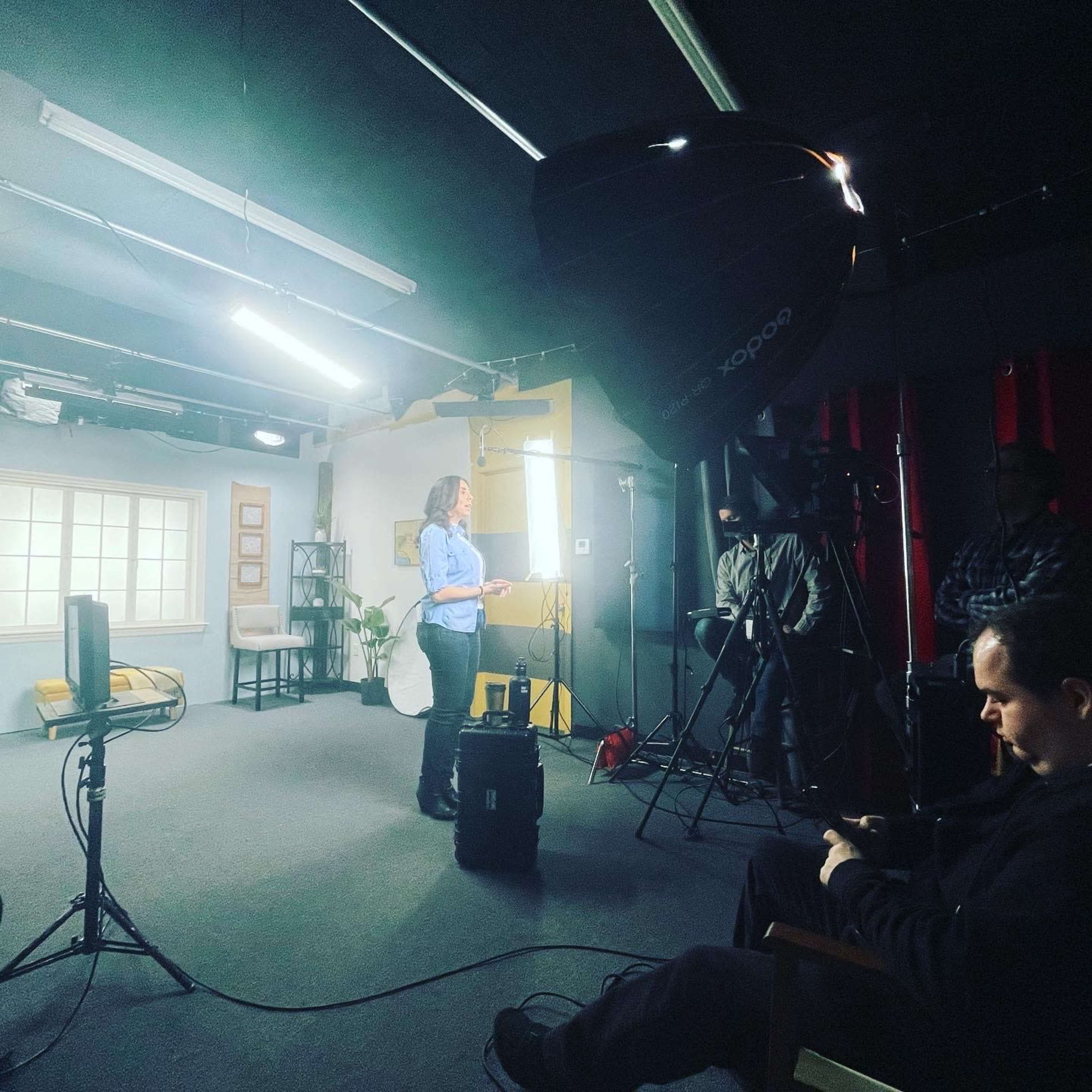 A woman stands in a well-lit studio, presenting while cameras and lights are set up around her.