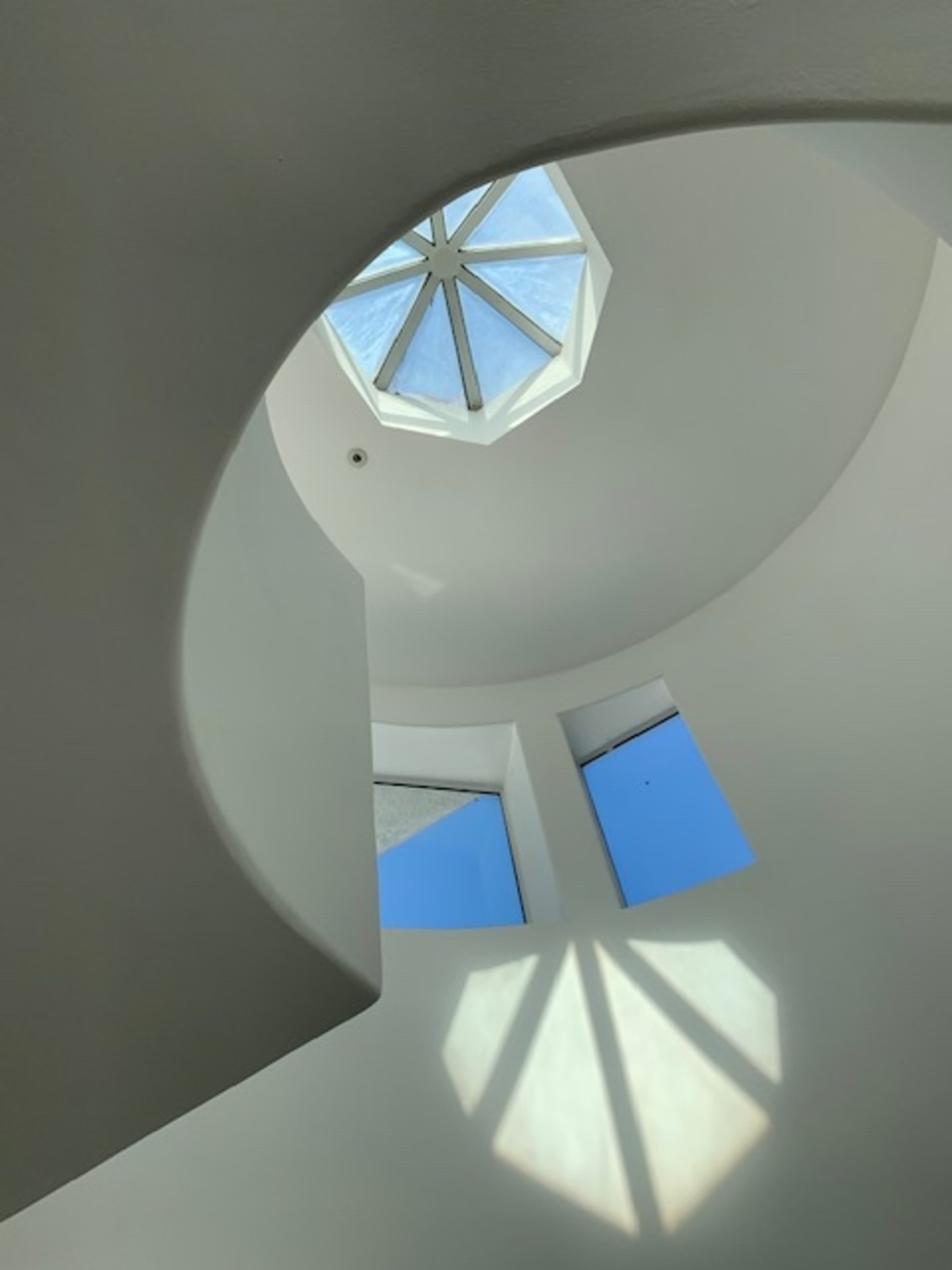 A circular stairwell with white walls, featuring a skylight and windows that allow blue sky to be visible.