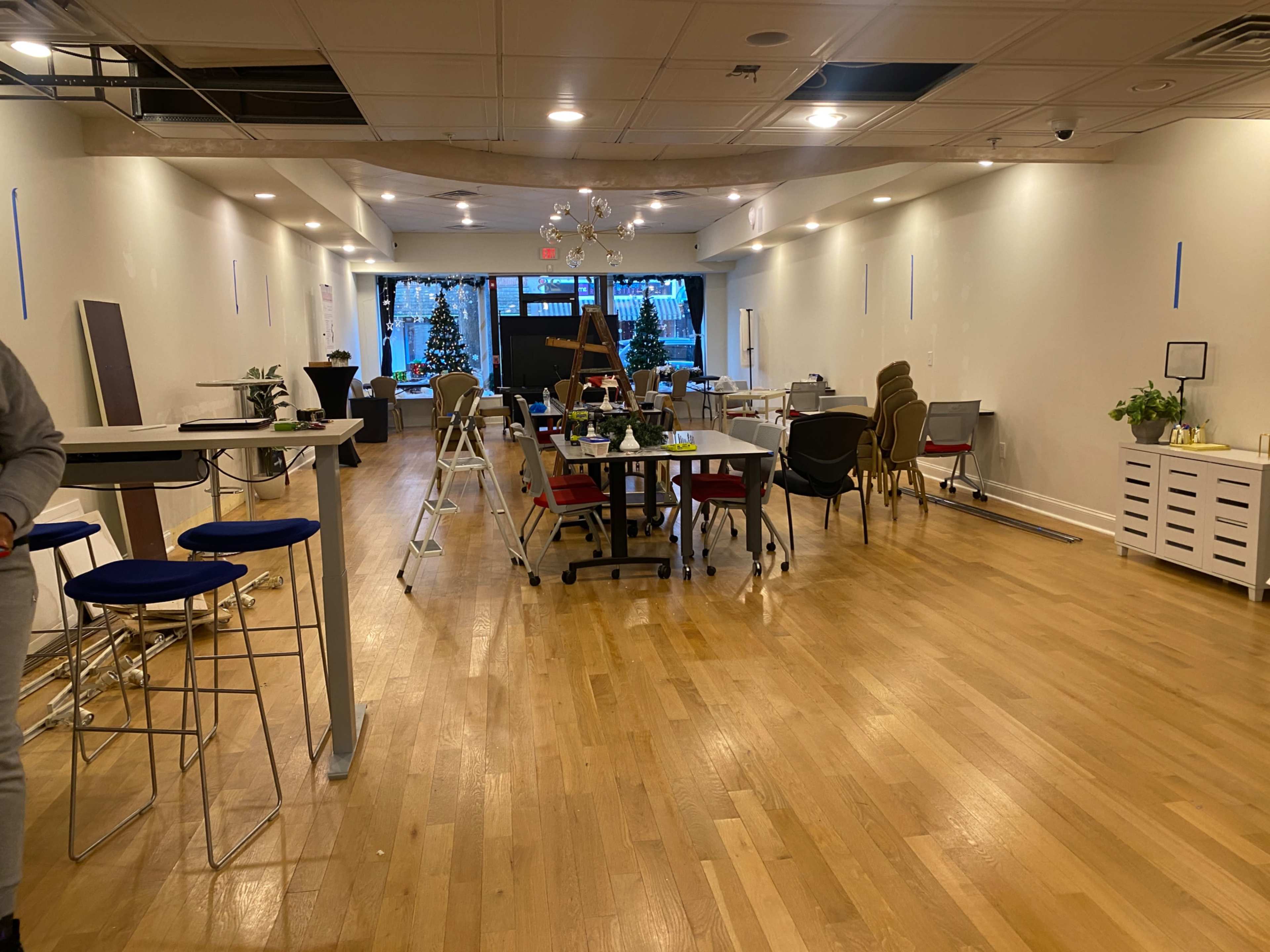 The image shows a spacious, partially furnished room with various tables, chairs, and a large window displaying a view of trees outside.
