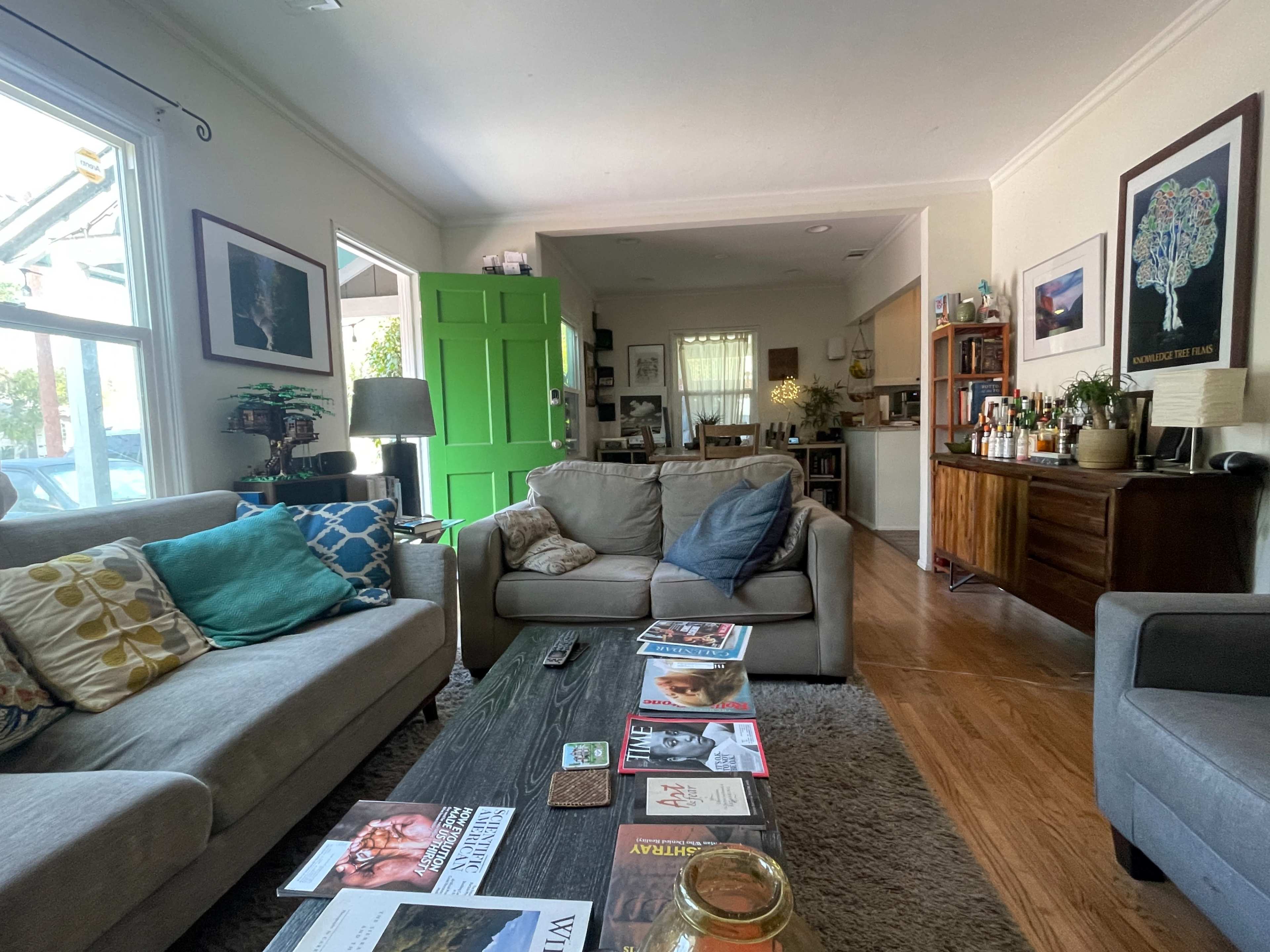 The living room features two sofas, a wooden coffee table with magazines, and a visible entrance to a kitchen area beyond a vibrant green door.