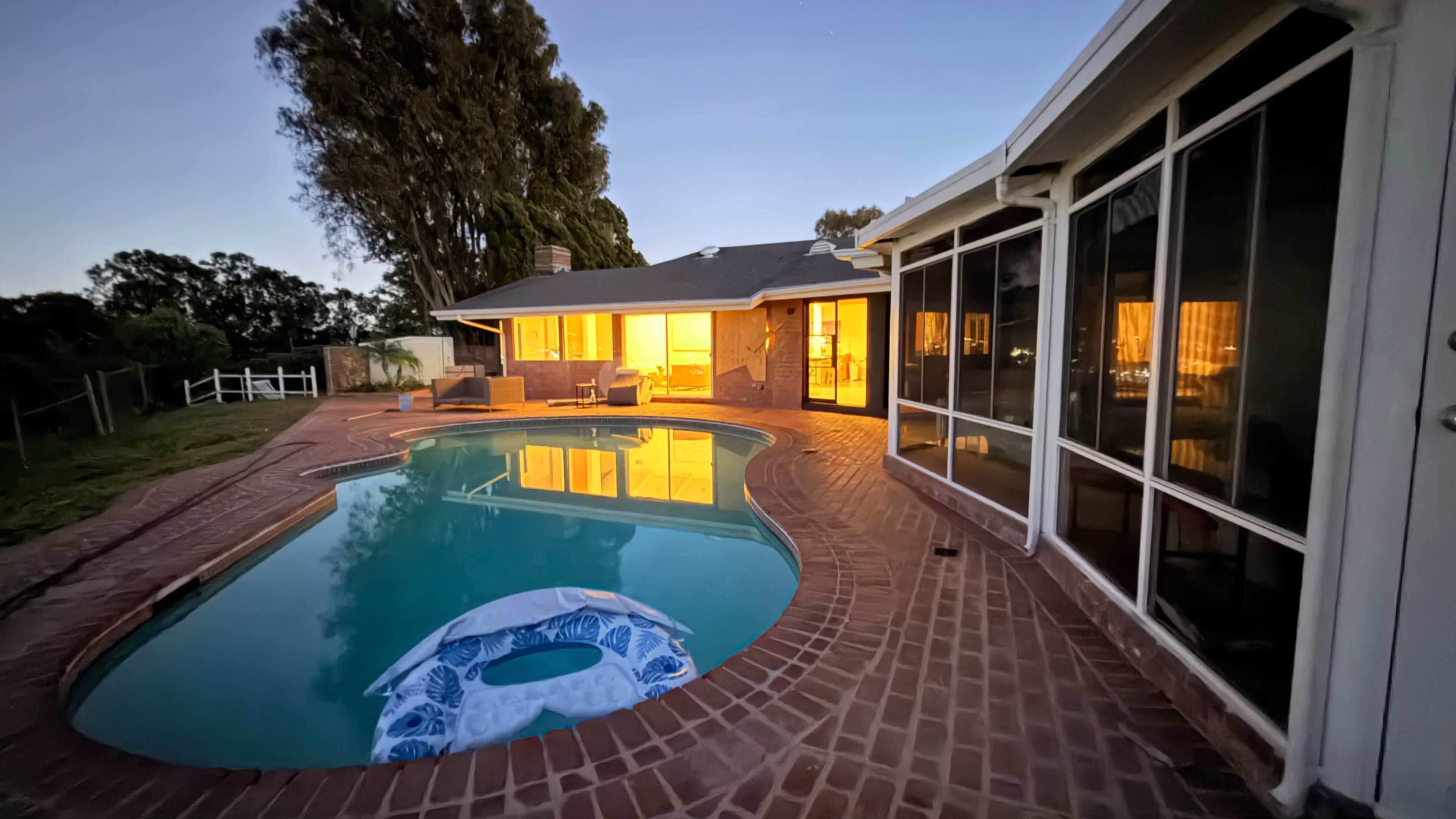 A still evening scene shows a swimming pool in front of a house, with warm light illuminating the interior and reflecting on the water's surface.