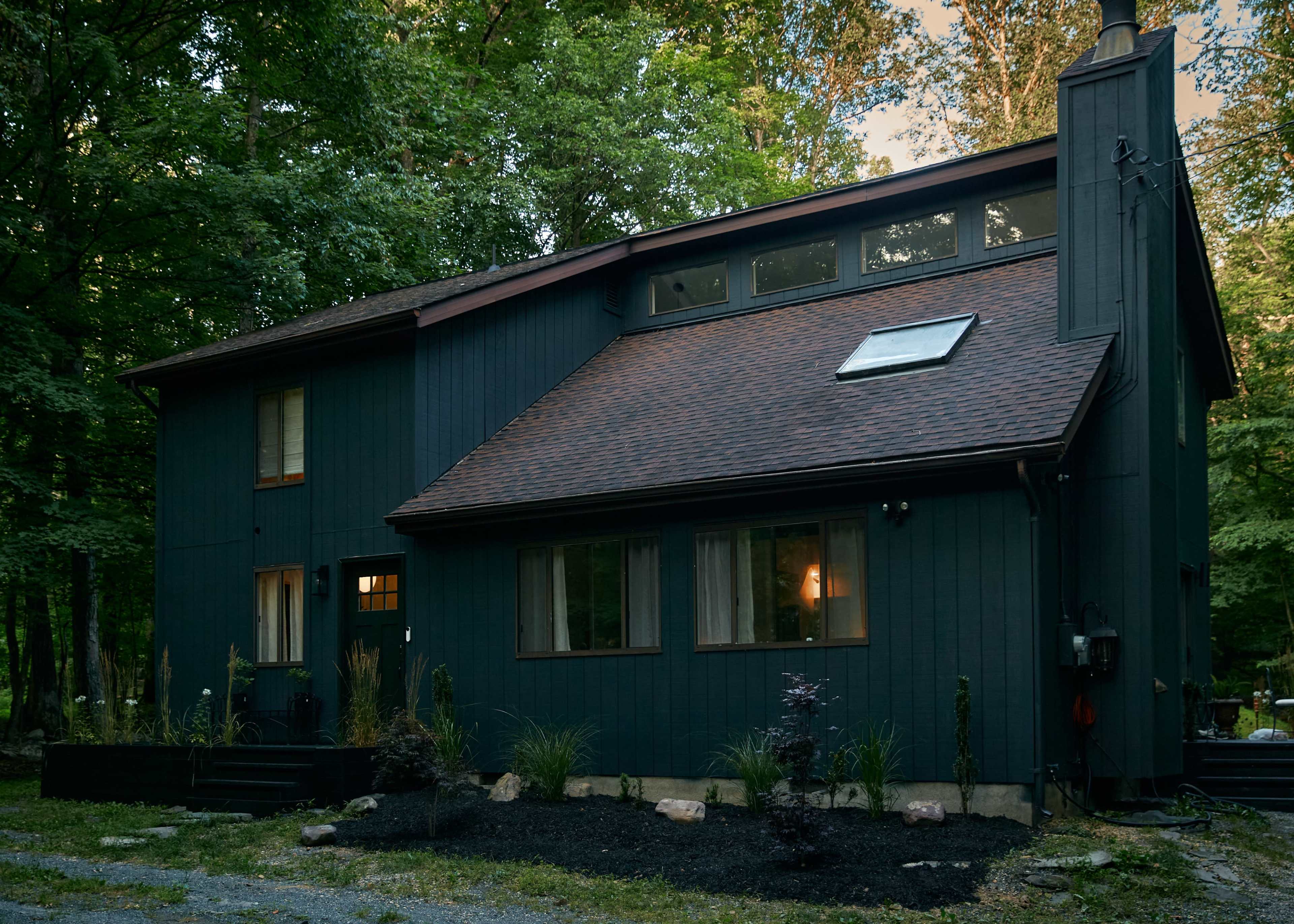 A two-story dark blue house with a sloped roof is surrounded by trees and features a landscaped garden with rocks and plants.
