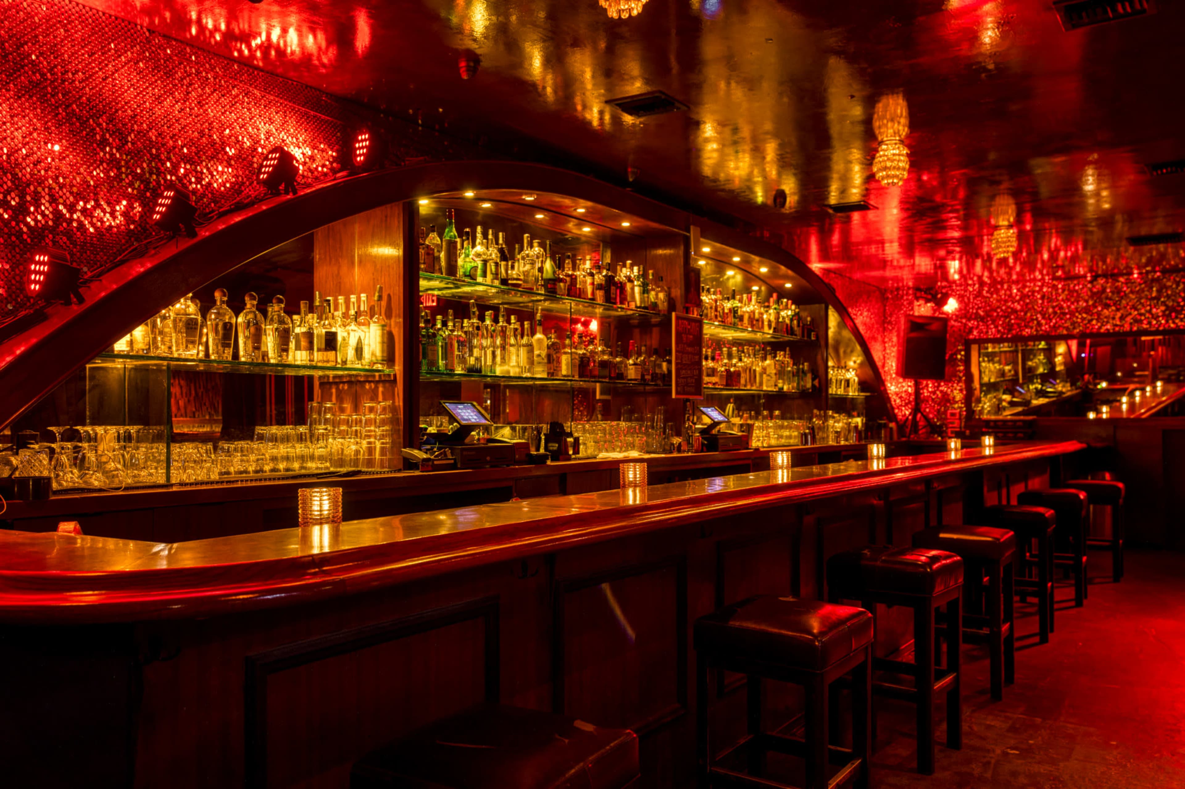 A dimly-lit bar with a curved backlit shelf filled with bottles and a long wooden counter with empty chairs.