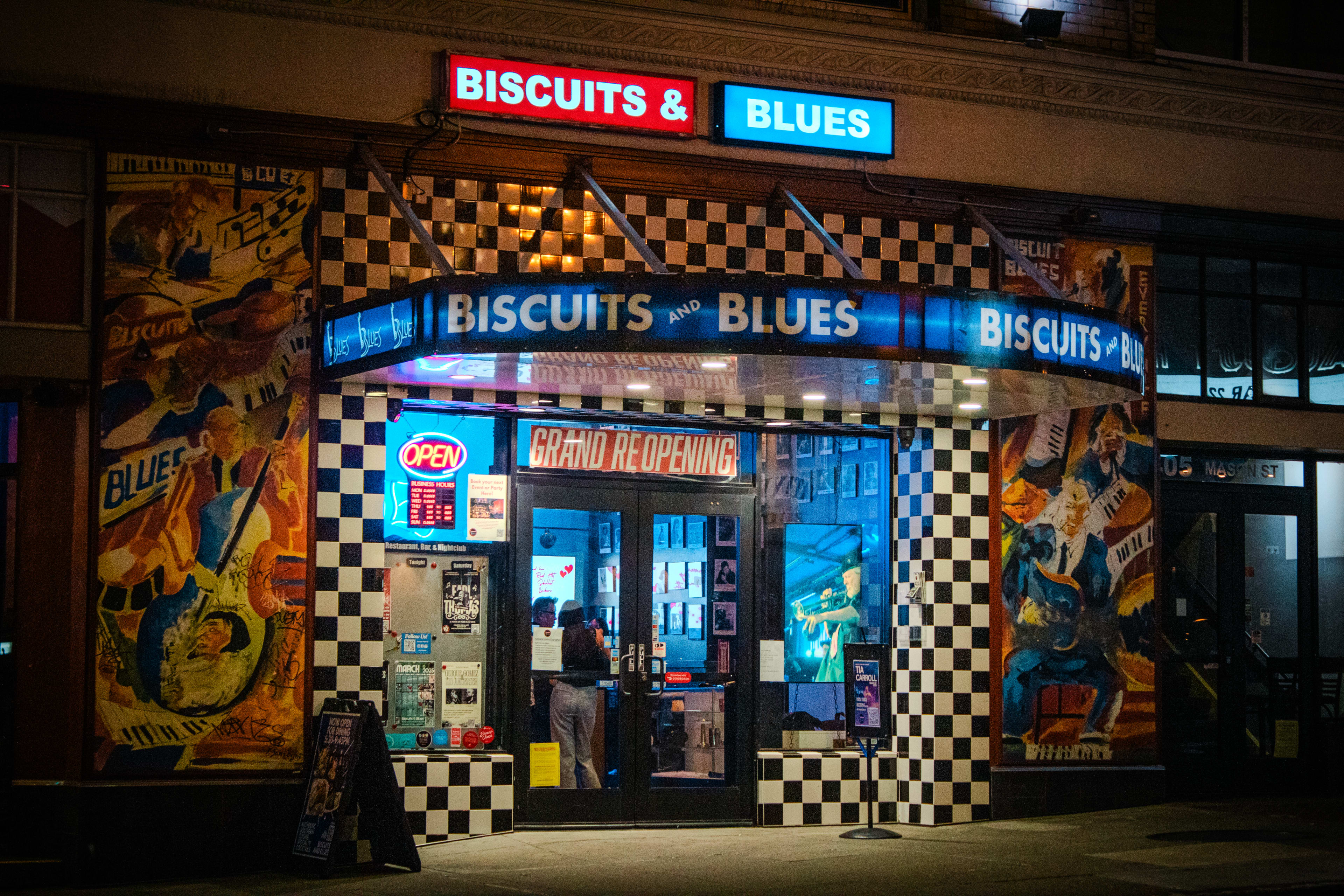 The image shows a nighttime view of a restaurant named "Biscuits & Blues," featuring a checkered exterior and colorful advertisements on the walls.