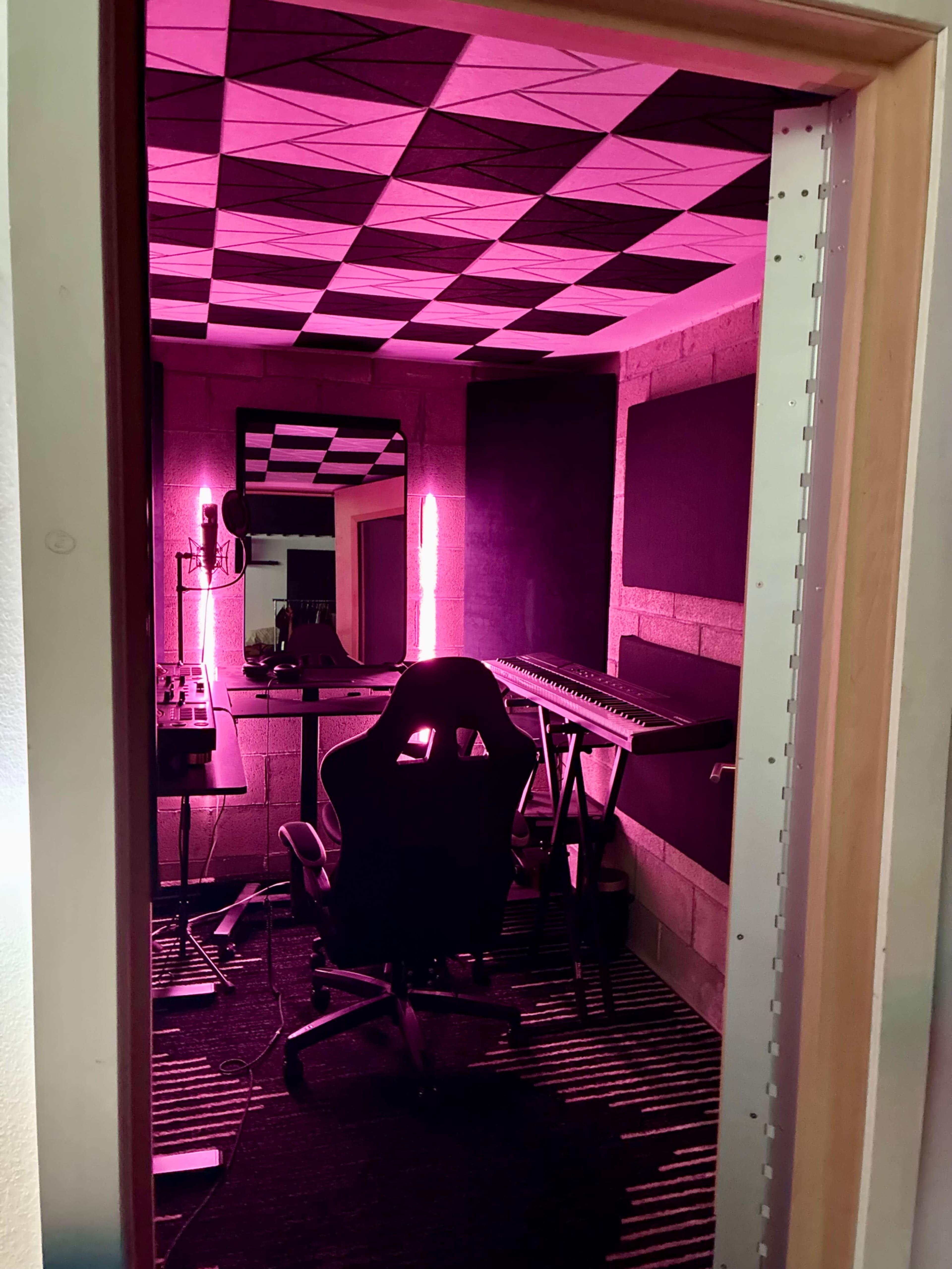 The image shows a music recording studio with pink lighting, featuring a desk, an office chair, and various musical equipment along the walls.
