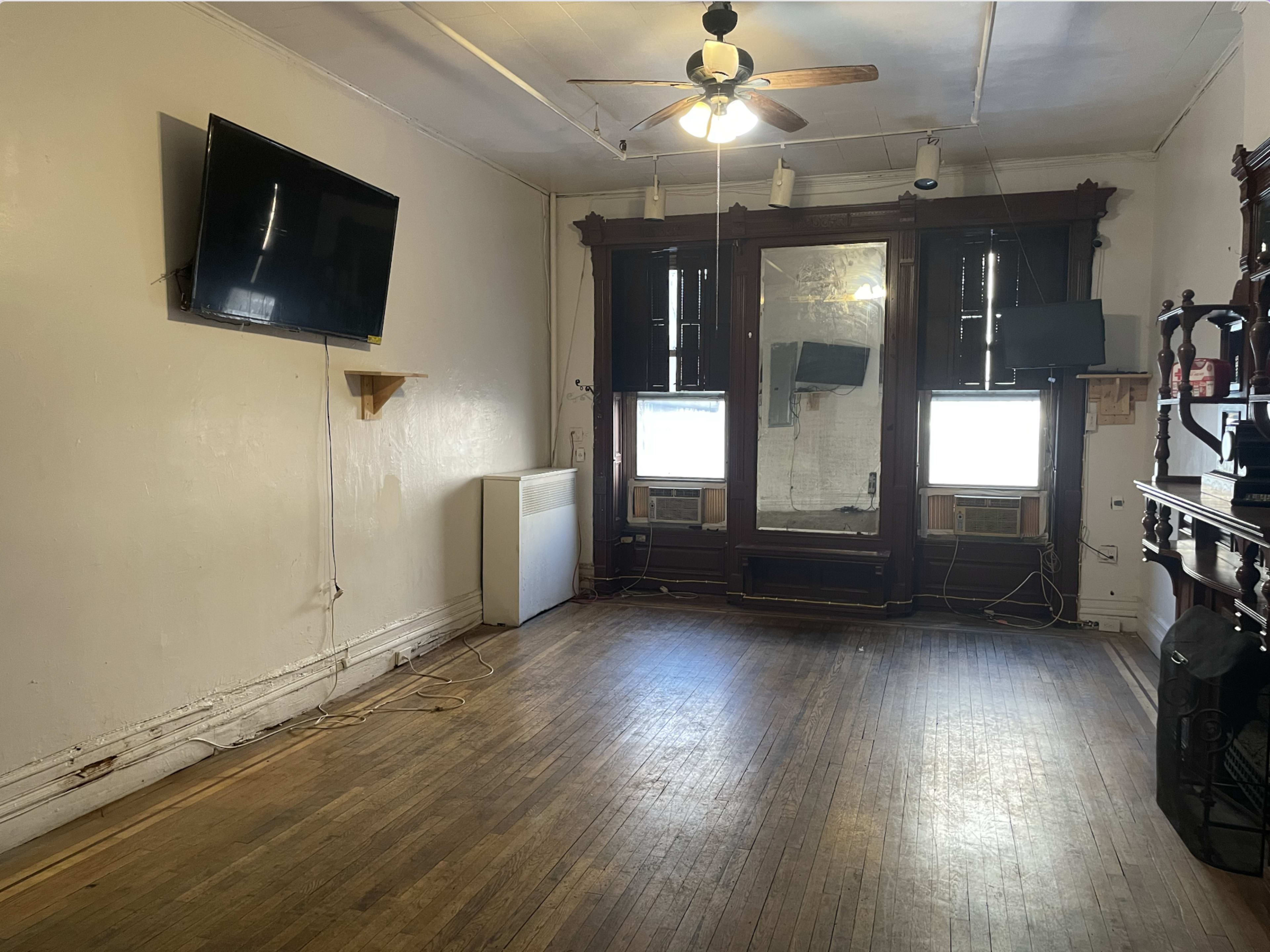 The image shows a sparsely furnished room with wooden floors, featuring a wall-mounted television, a ceiling fan, and two windows with air conditioning units.