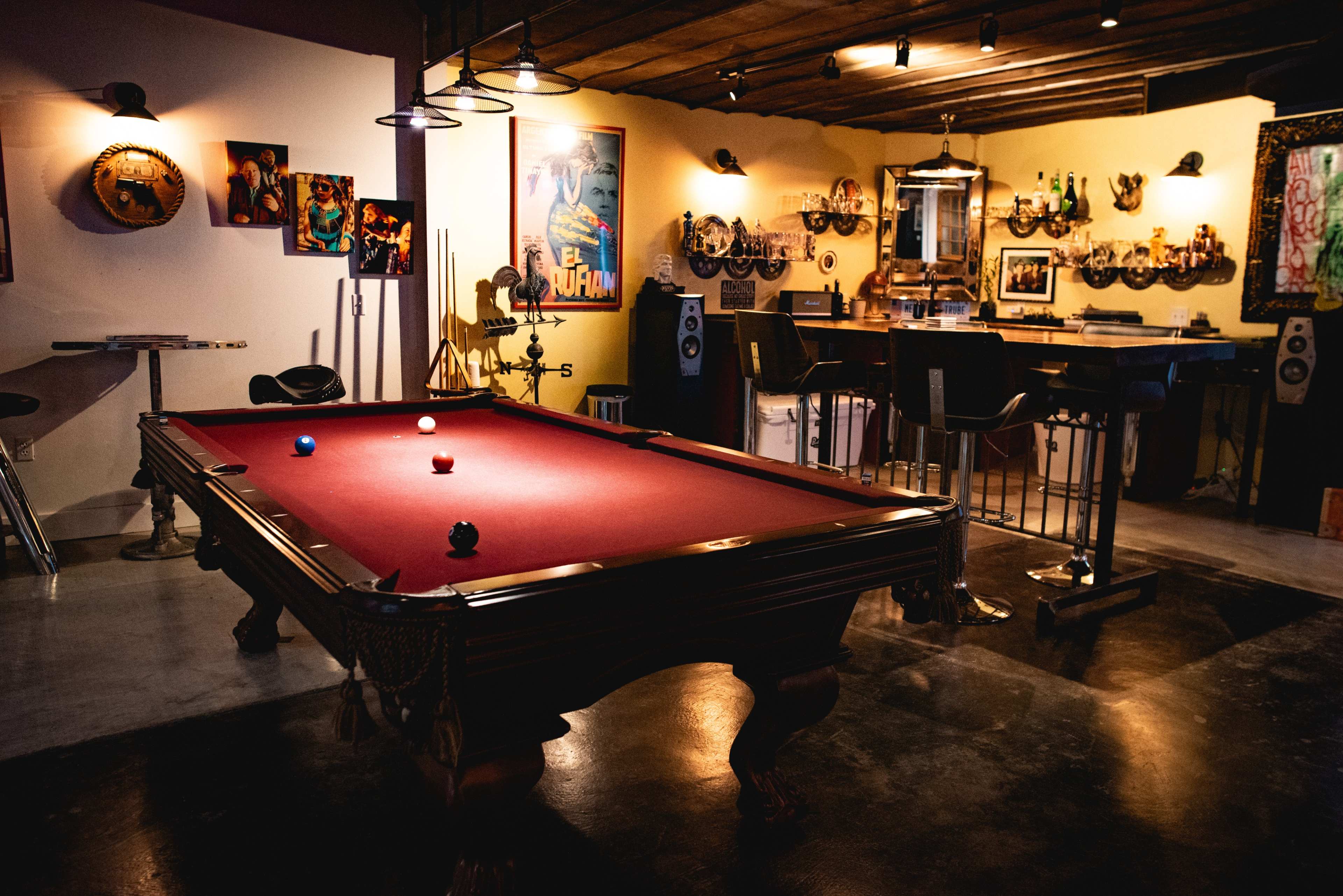 A game room features a red pool table with three balls positioned on it, surrounded by framed artwork and bar seating in a well-lit space.