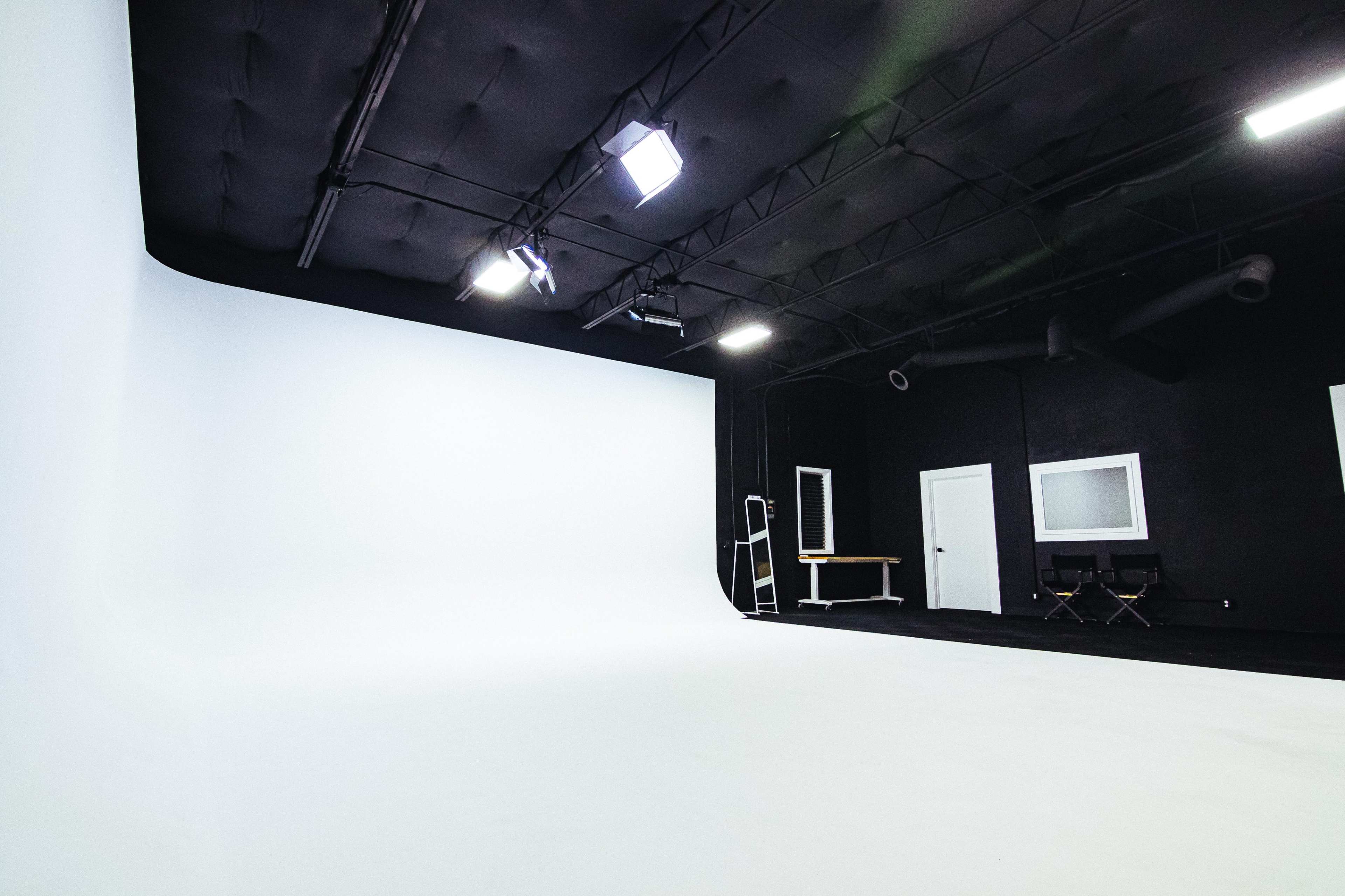 The image shows a photography studio with a white backdrop, black walls, and a few lighting fixtures.