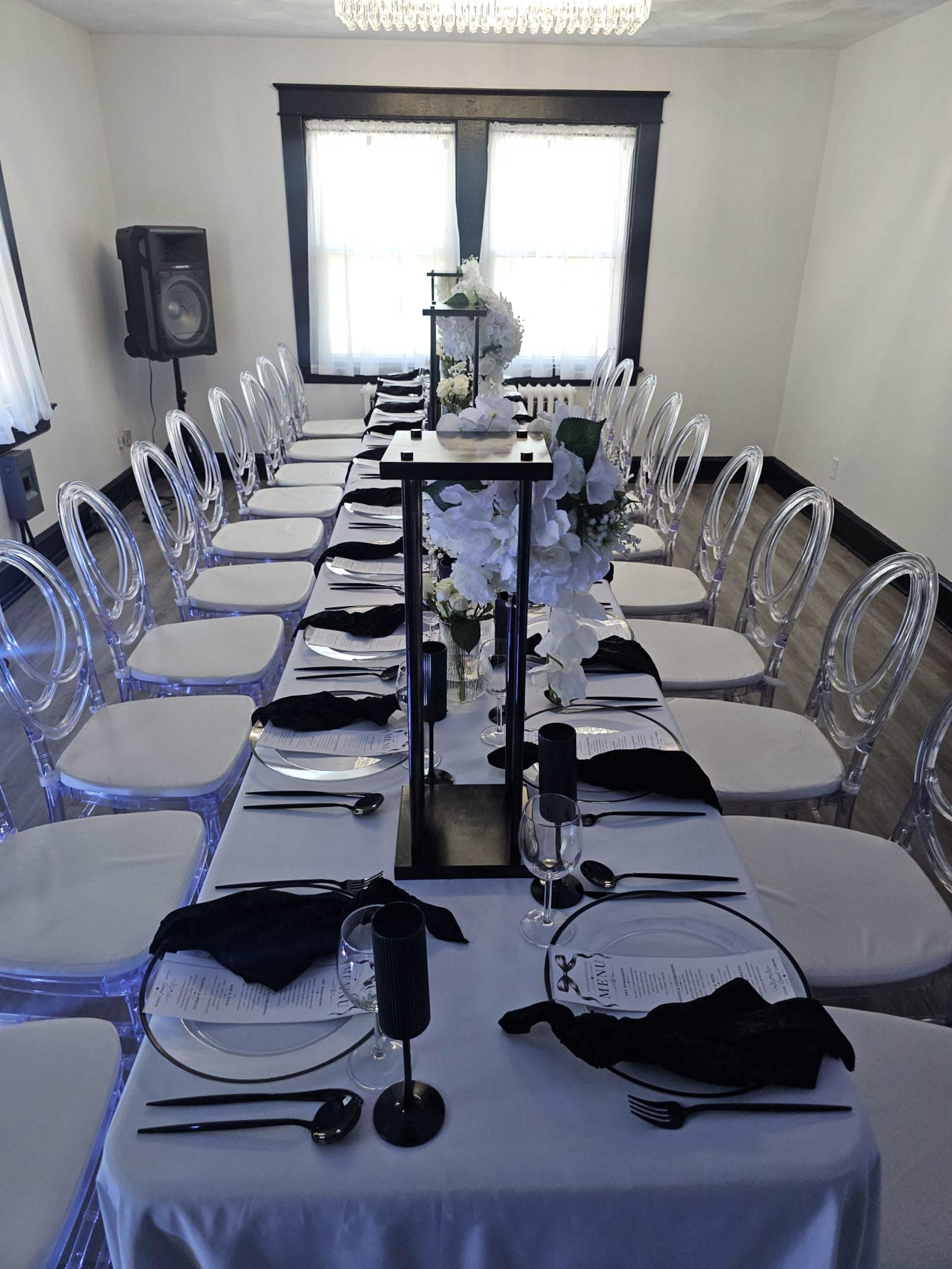 A rectangular banquet table is set with clear chairs, black napkins, silverware, and elegant centerpieces in a well-lit room.