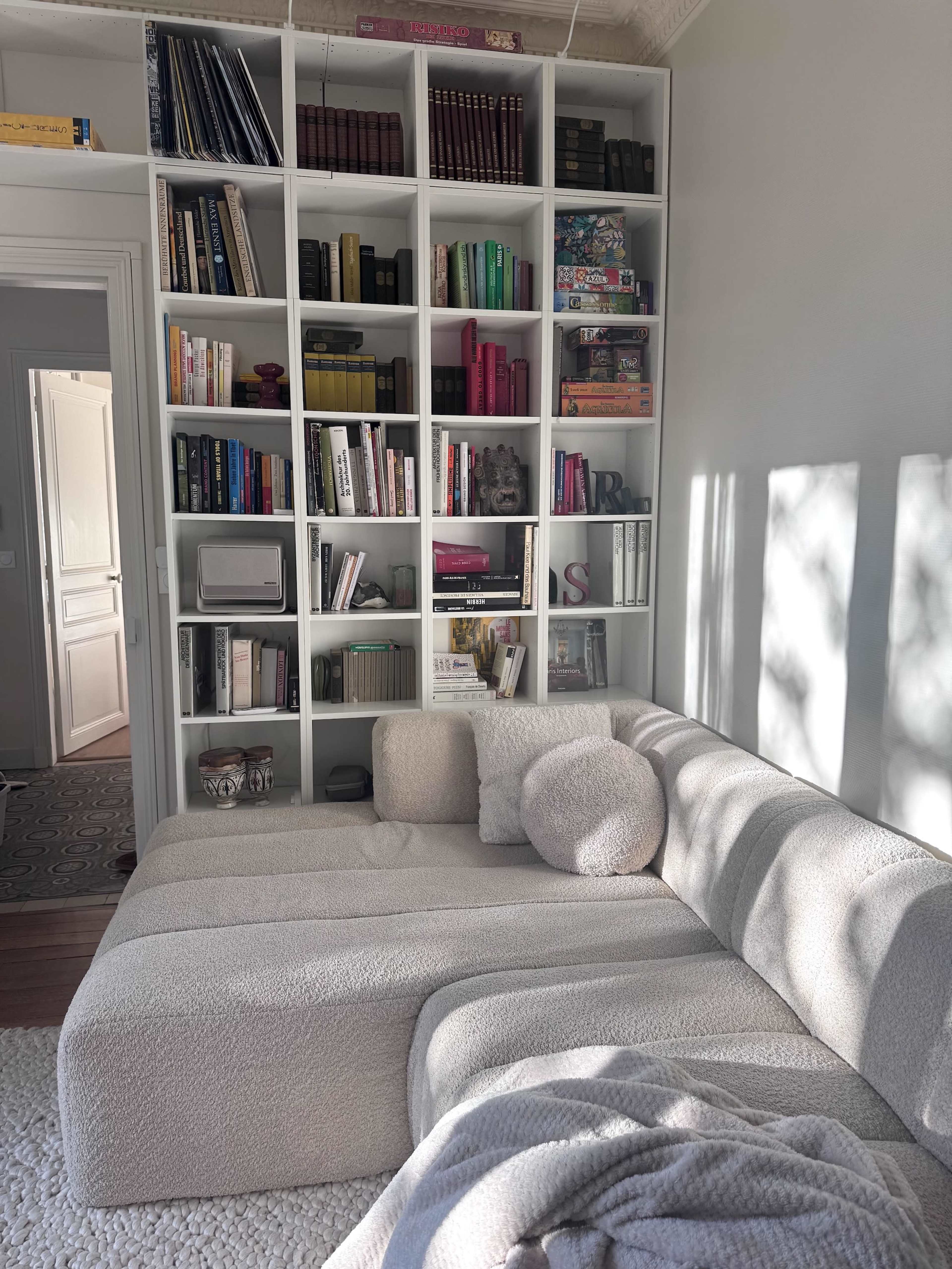 A cozy living room features a light-colored, sectional sofa and a tall bookshelf filled with books and decorative items.
